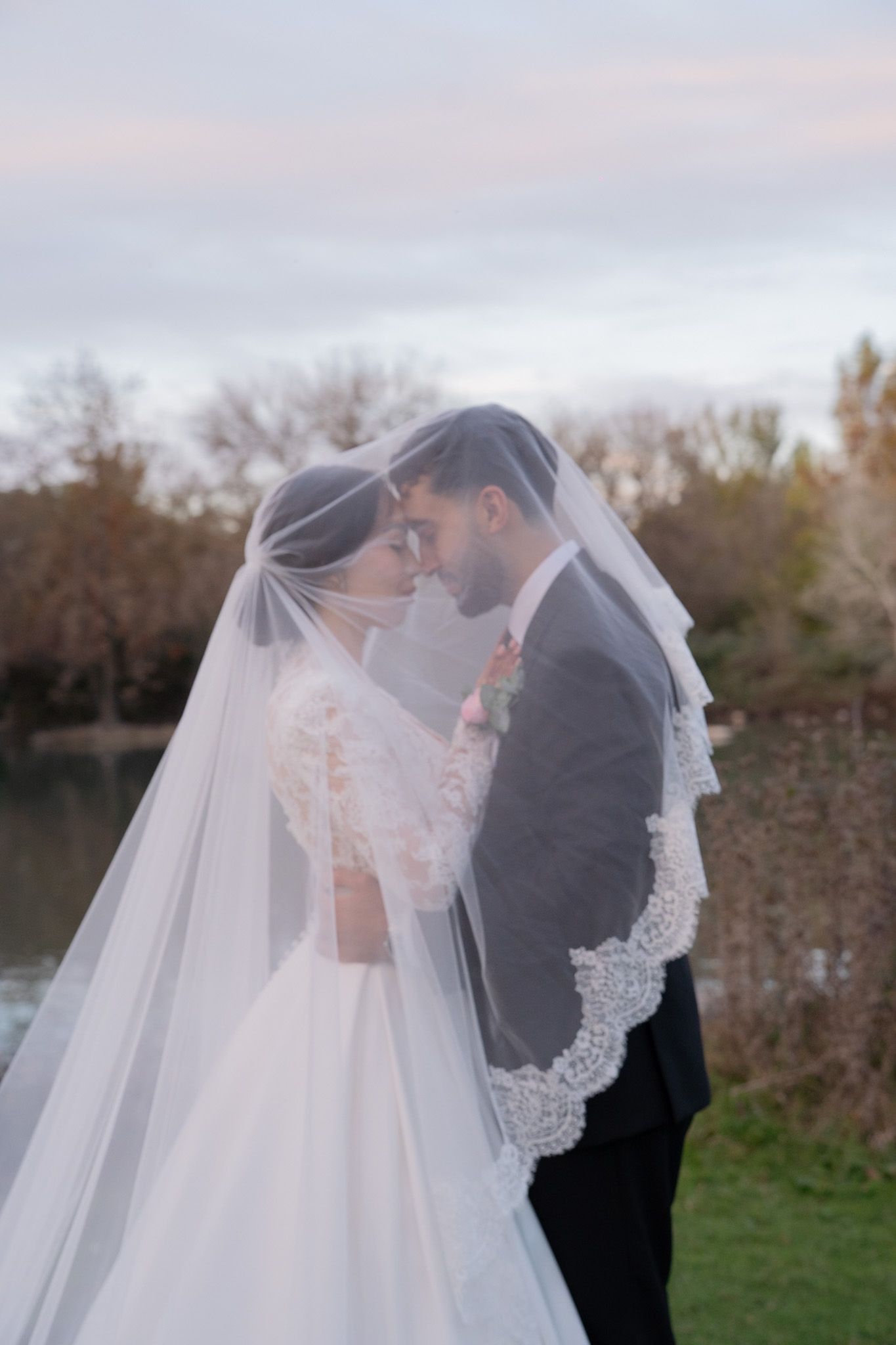 Bride and groom share romantic moment under lace-trimmed wedding veil outdoors near lake at sunset.