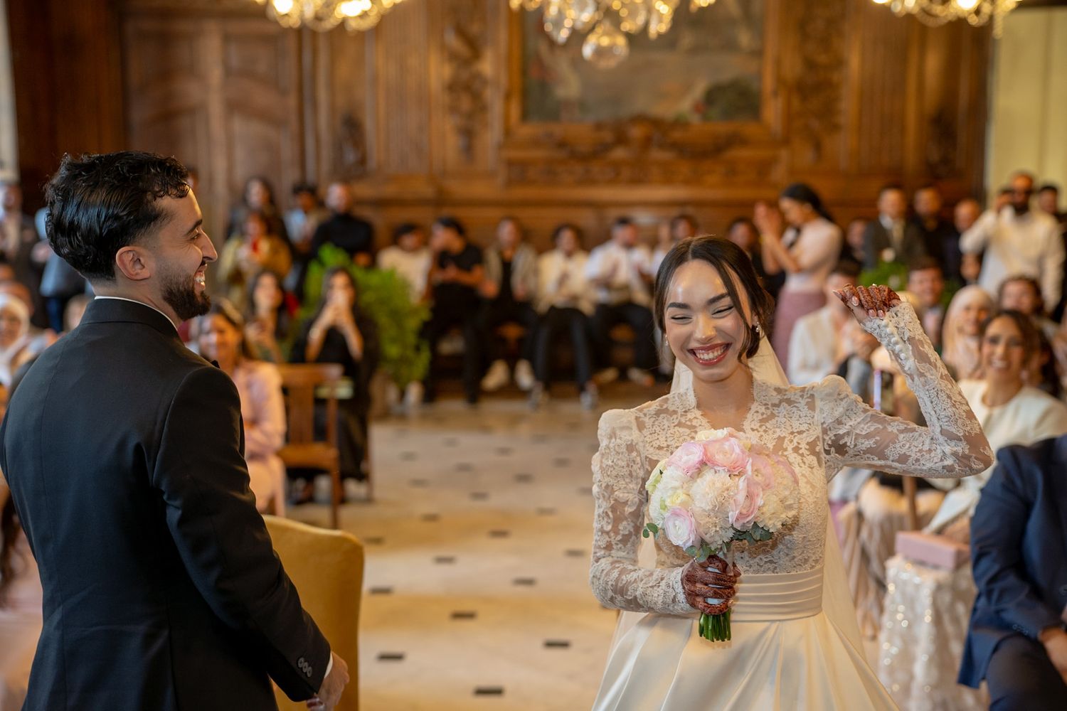 Bride smiling joyfully holding a bouquet during an elegant indoor wedding ceremony with guests seated in the background.