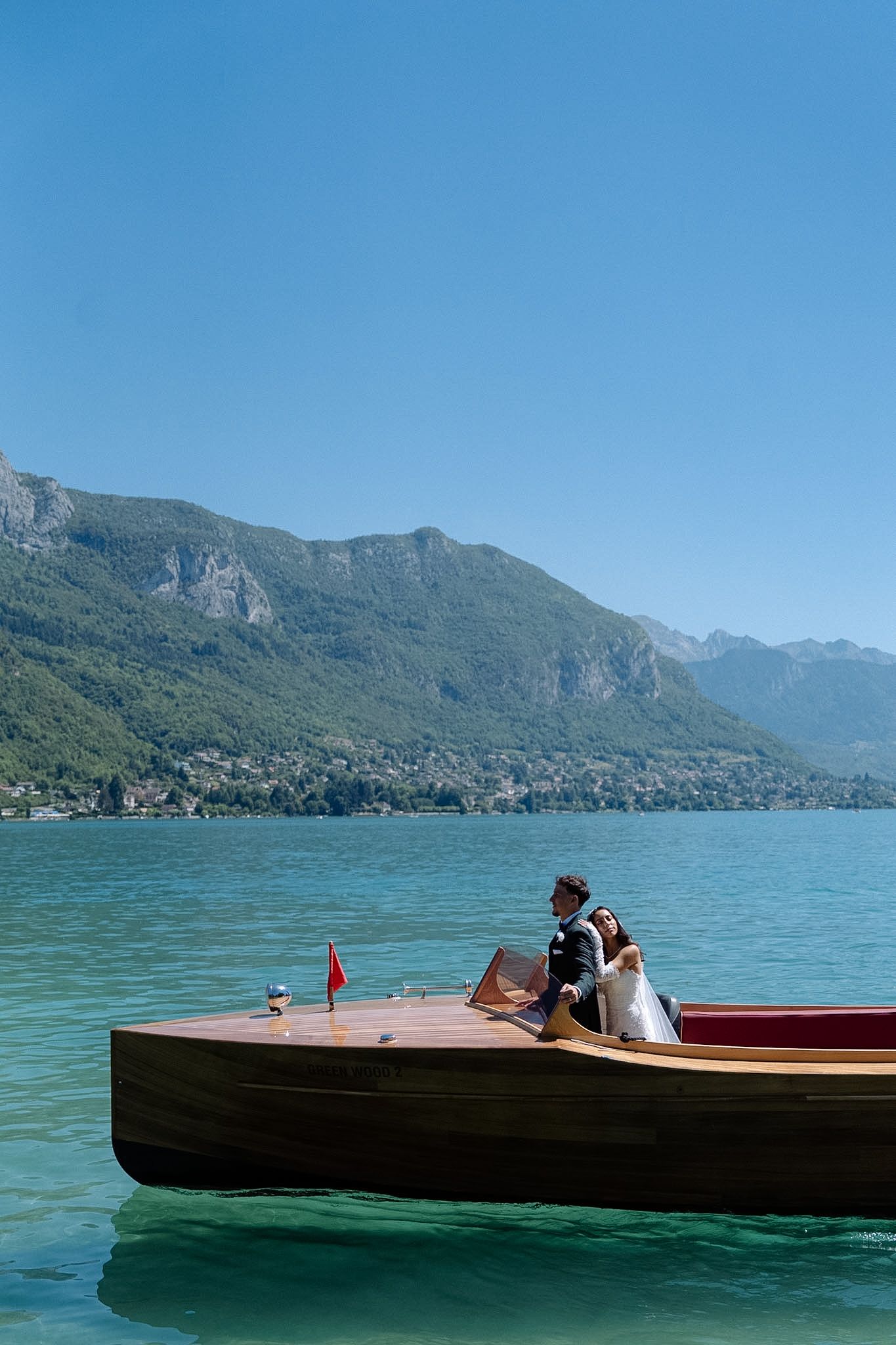 Bride and groom sitting on a classic wooden boat on a turquoise alpine lake with mountain backdrop.