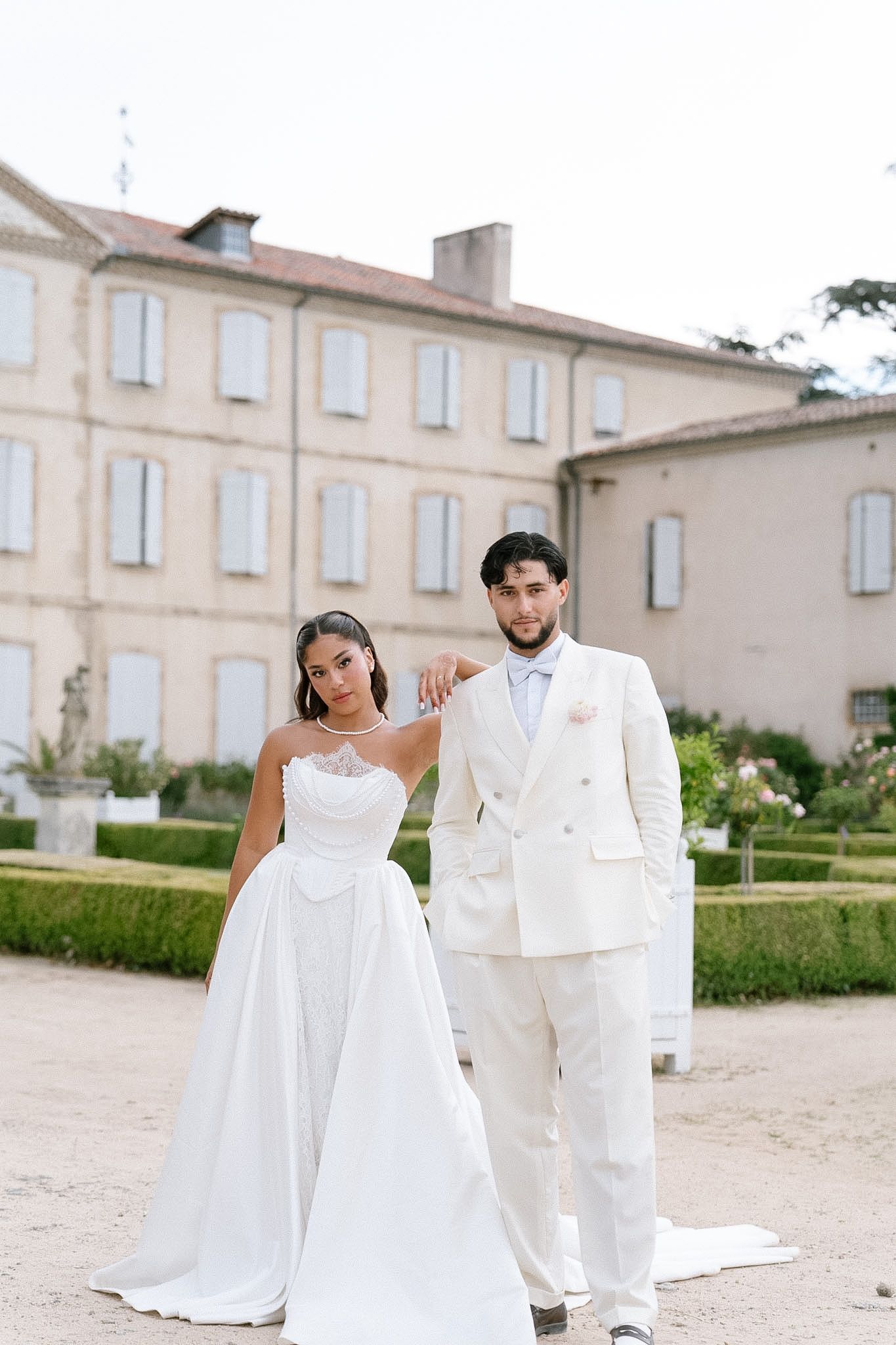 Bride in white strapless gown and groom in cream suit pose together outside a French château.