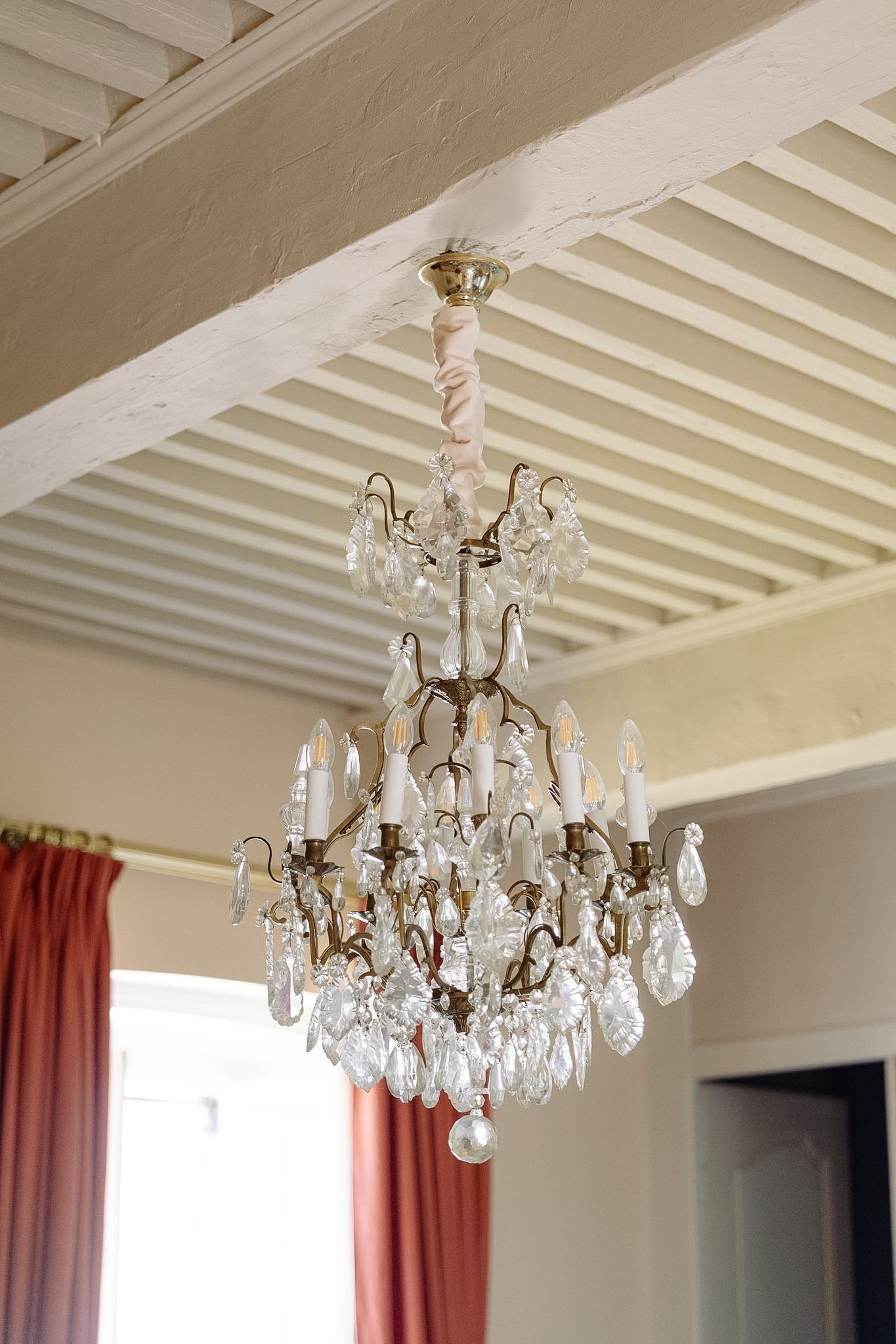Elegant crystal chandelier with candle lights hanging from white beamed ceiling in a classic French interior.