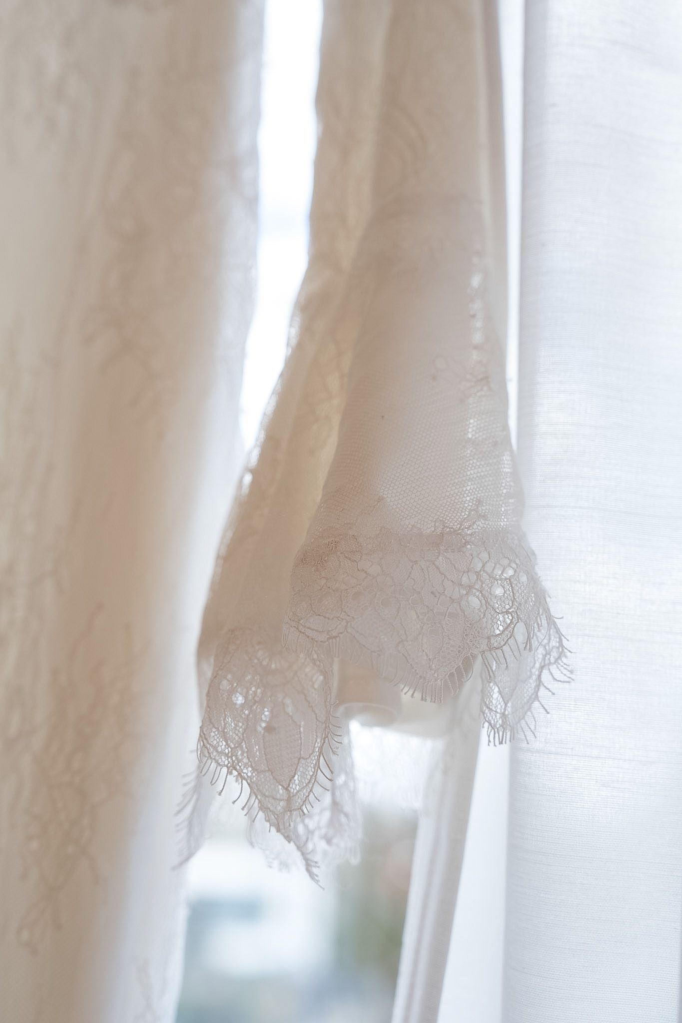 Close-up of delicate white lace trim on a flowing bridal gown hanging near a bright window.