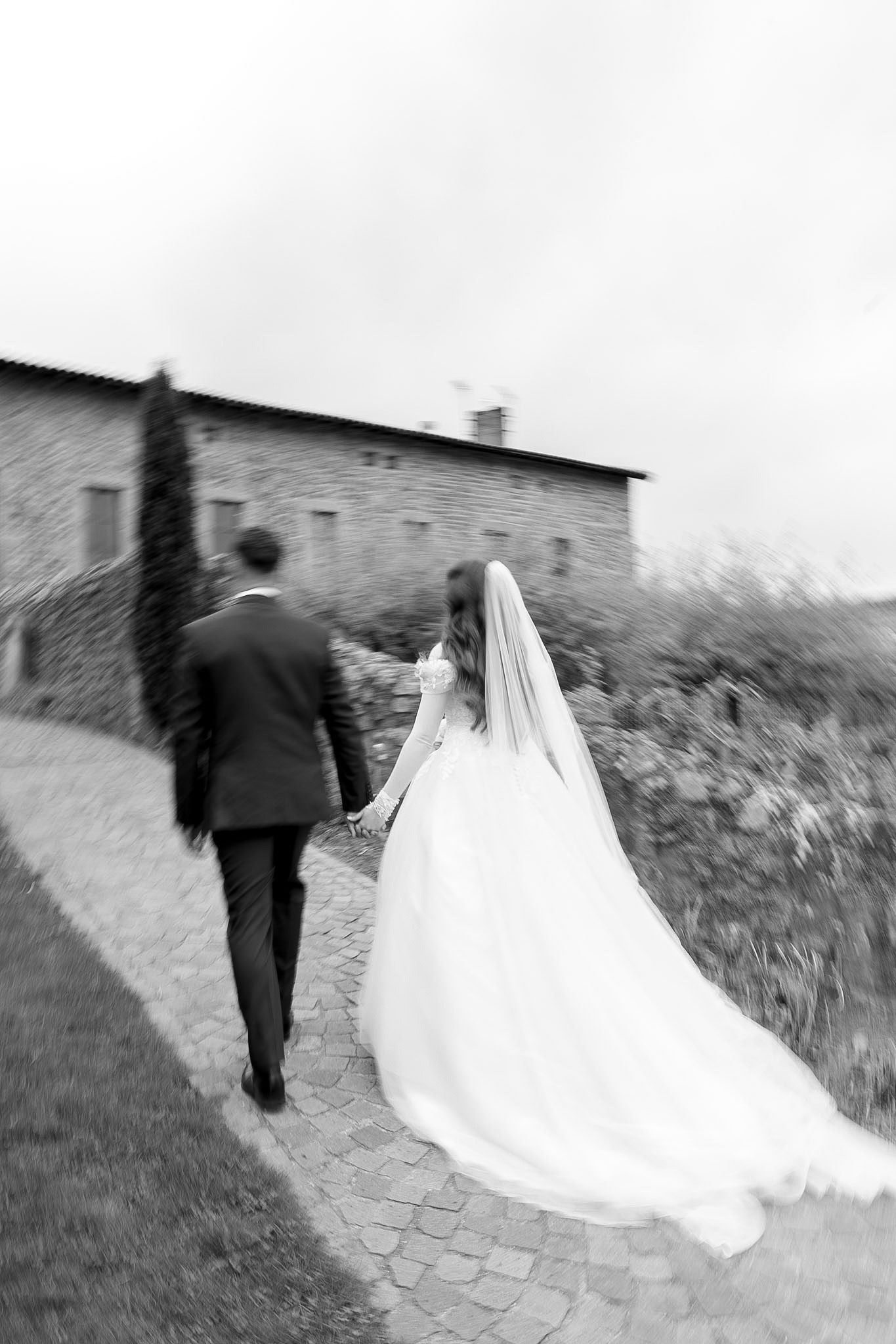 Black and white photo of bride and groom walking hand in hand along a cobblestone path toward a rustic stone building.