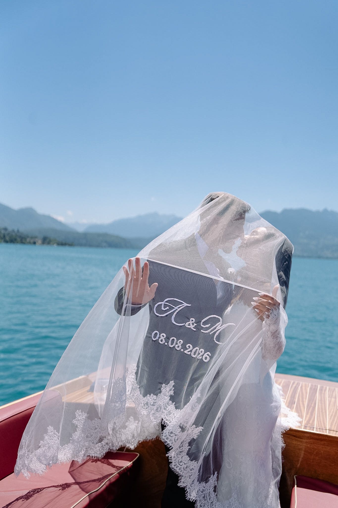 Bride and groom kissing behind a wedding sign on a boat, with turquoise lake and mountains in background.