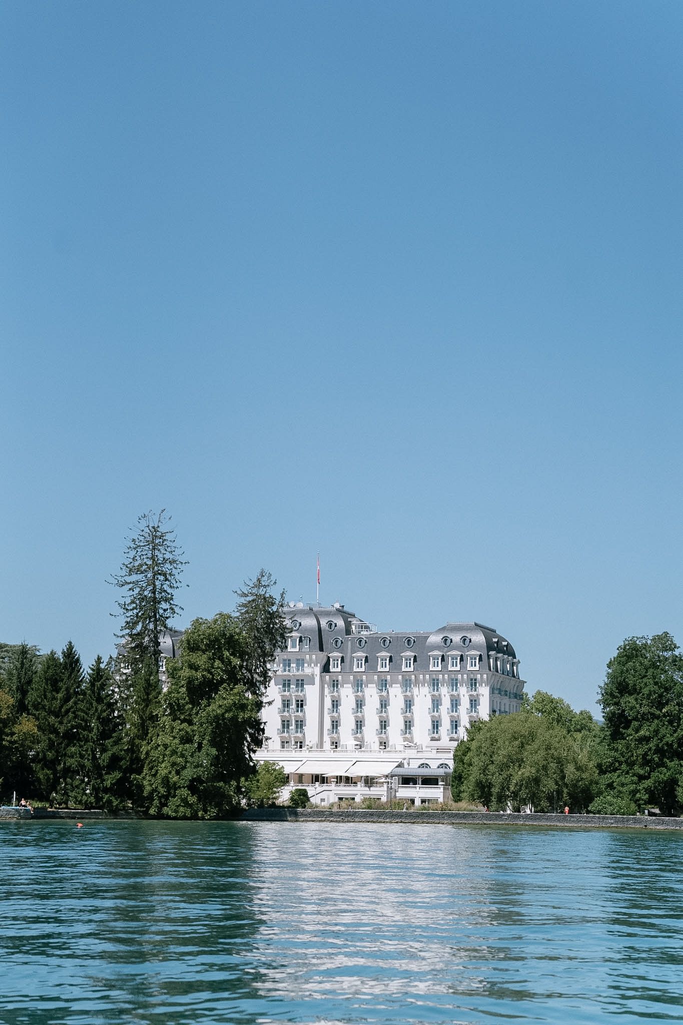 Grand white hotel beside a calm lake surrounded by lush green trees under a clear blue sky.