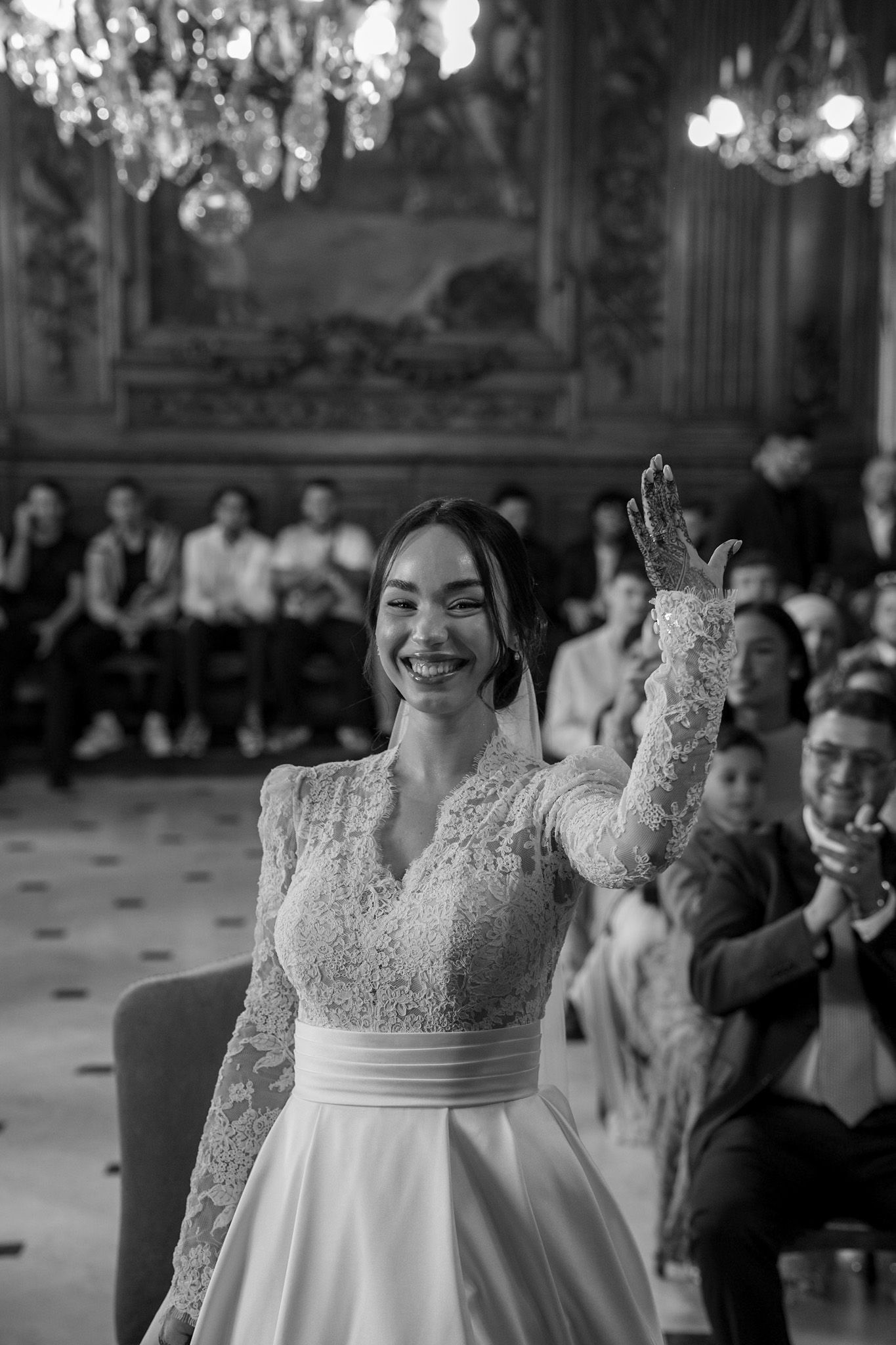 Joyful bride in lace gown celebrating at wedding ceremony in ornate chandelier ballroom.