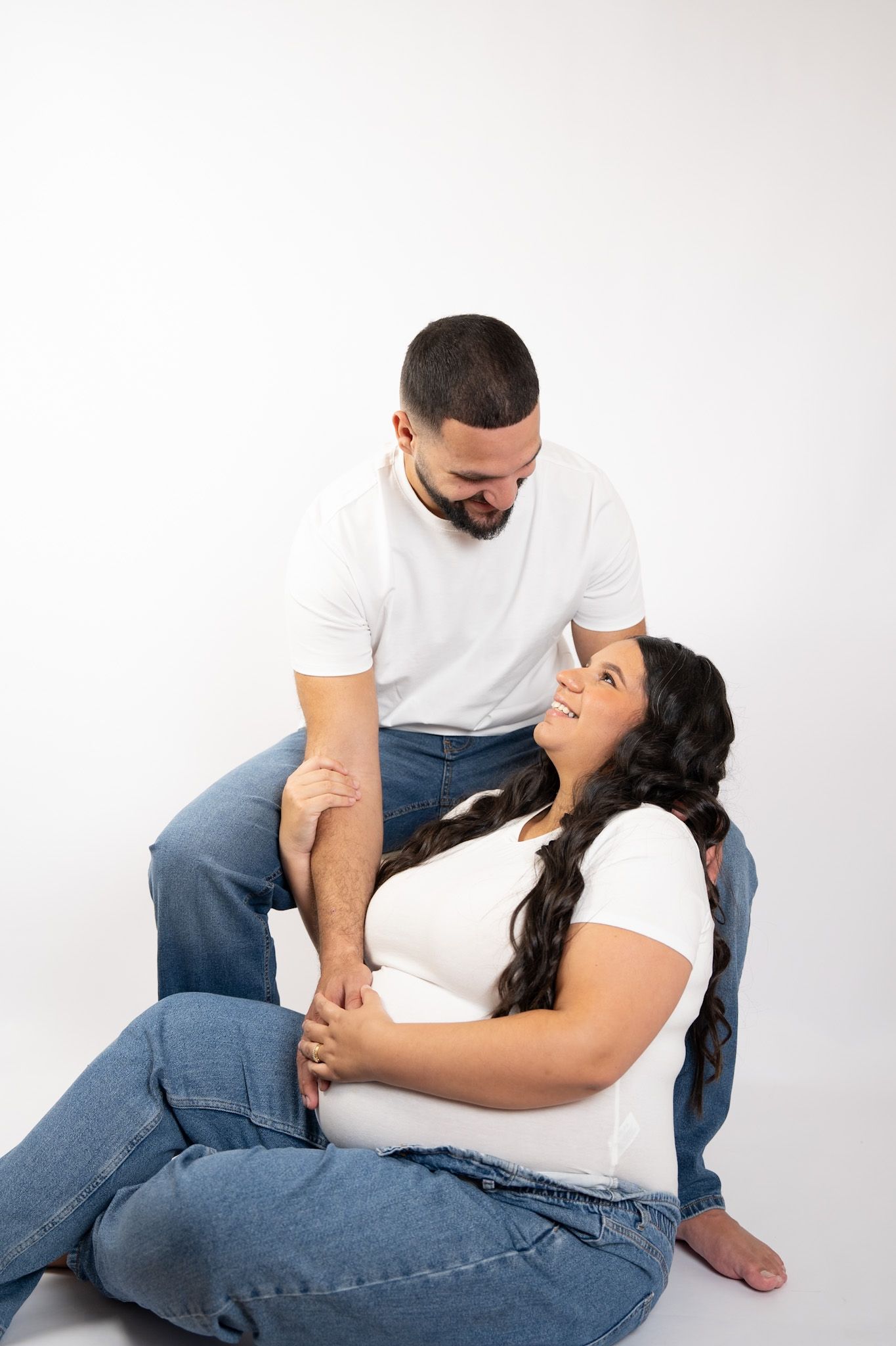 Pregnant couple in white shirts and jeans posing together, woman sitting on floor smiling up at partner.