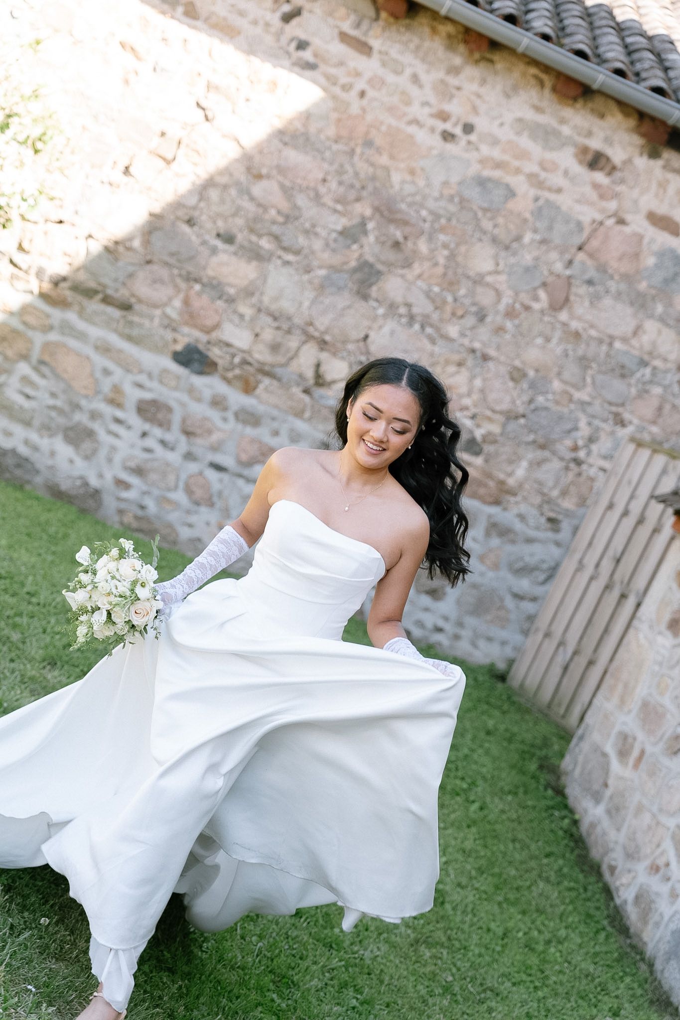 Smiling bride in strapless white gown with gloves holds bouquet outdoors near rustic stone wall.
