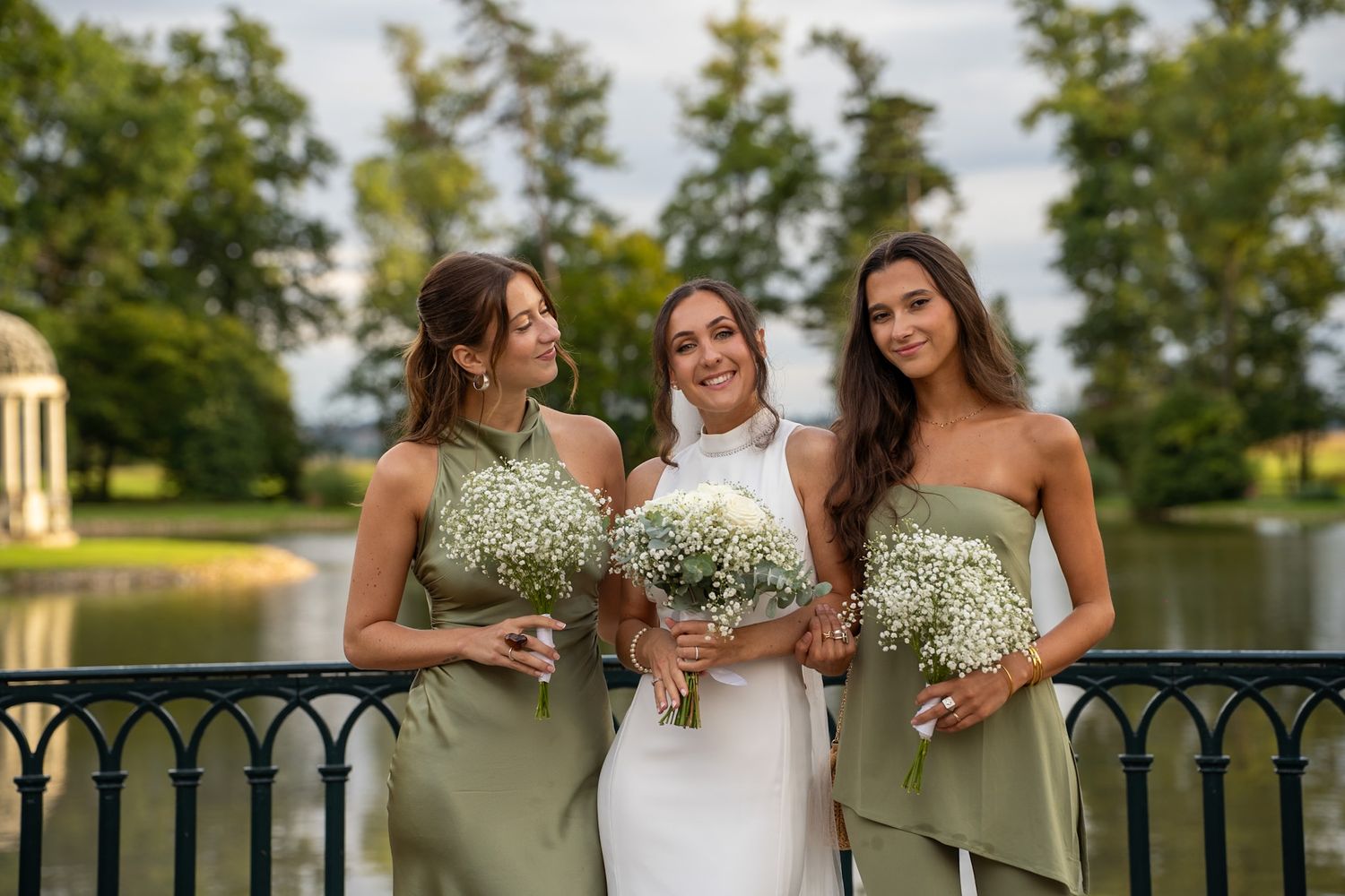 Bride in white gown with two bridesmaids in sage green dresses holding baby's breath bouquets on a bridge.