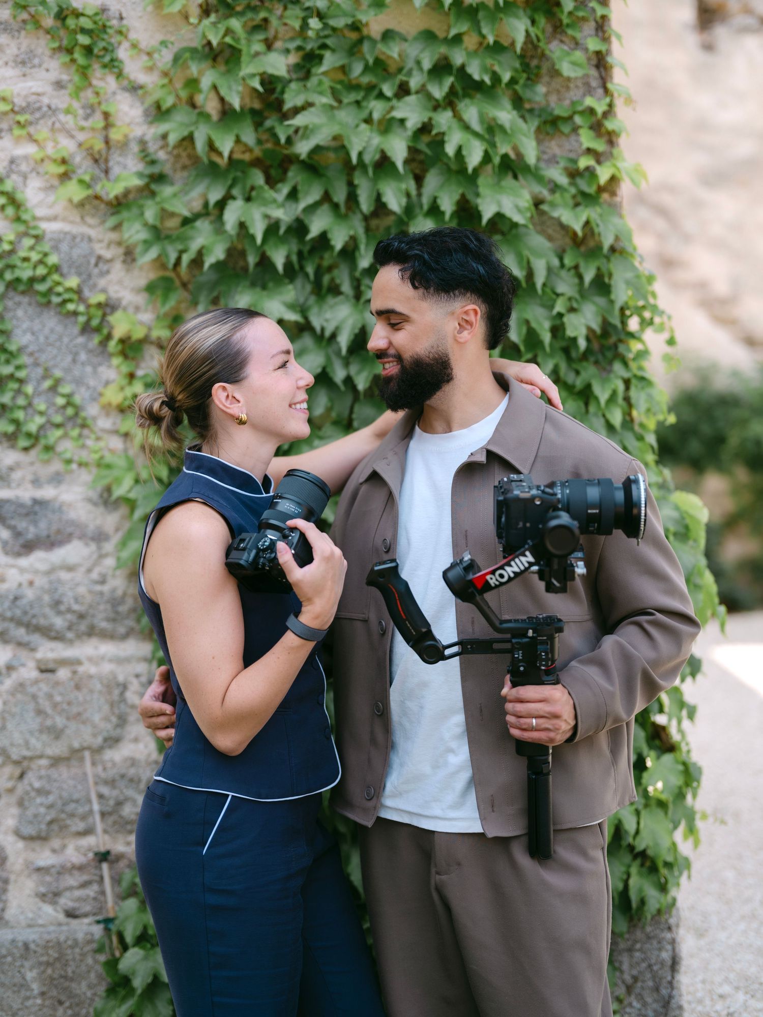 A couple holding cameras and a gimbal smiling at each other outdoors against an ivy-covered stone wall.