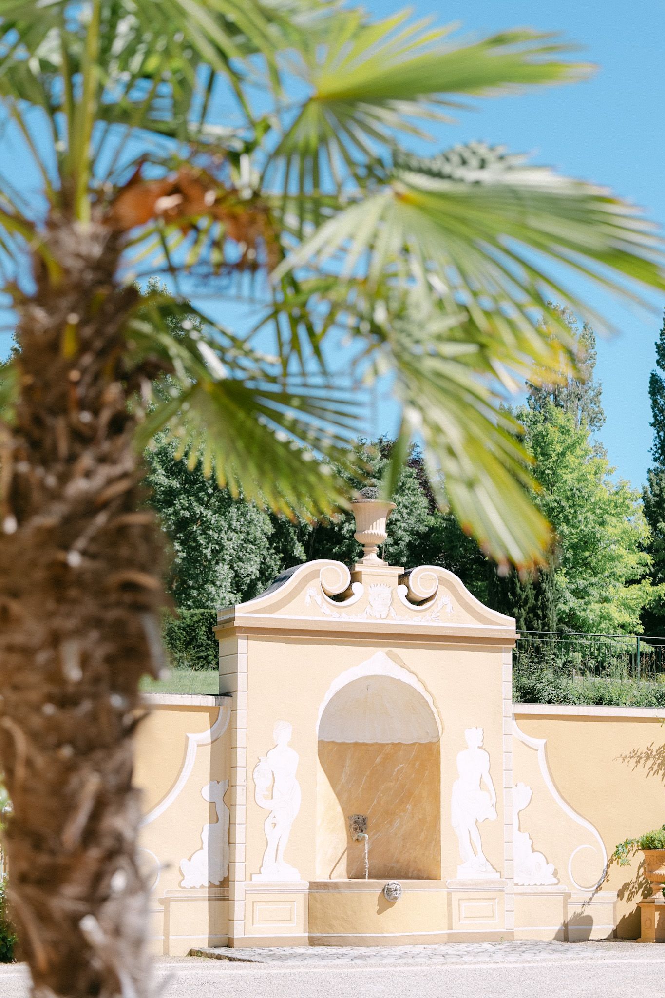 Ornate yellow stone fountain with classical sculptures set against lush green trees and blue sky, framed by a tropical palm tree.