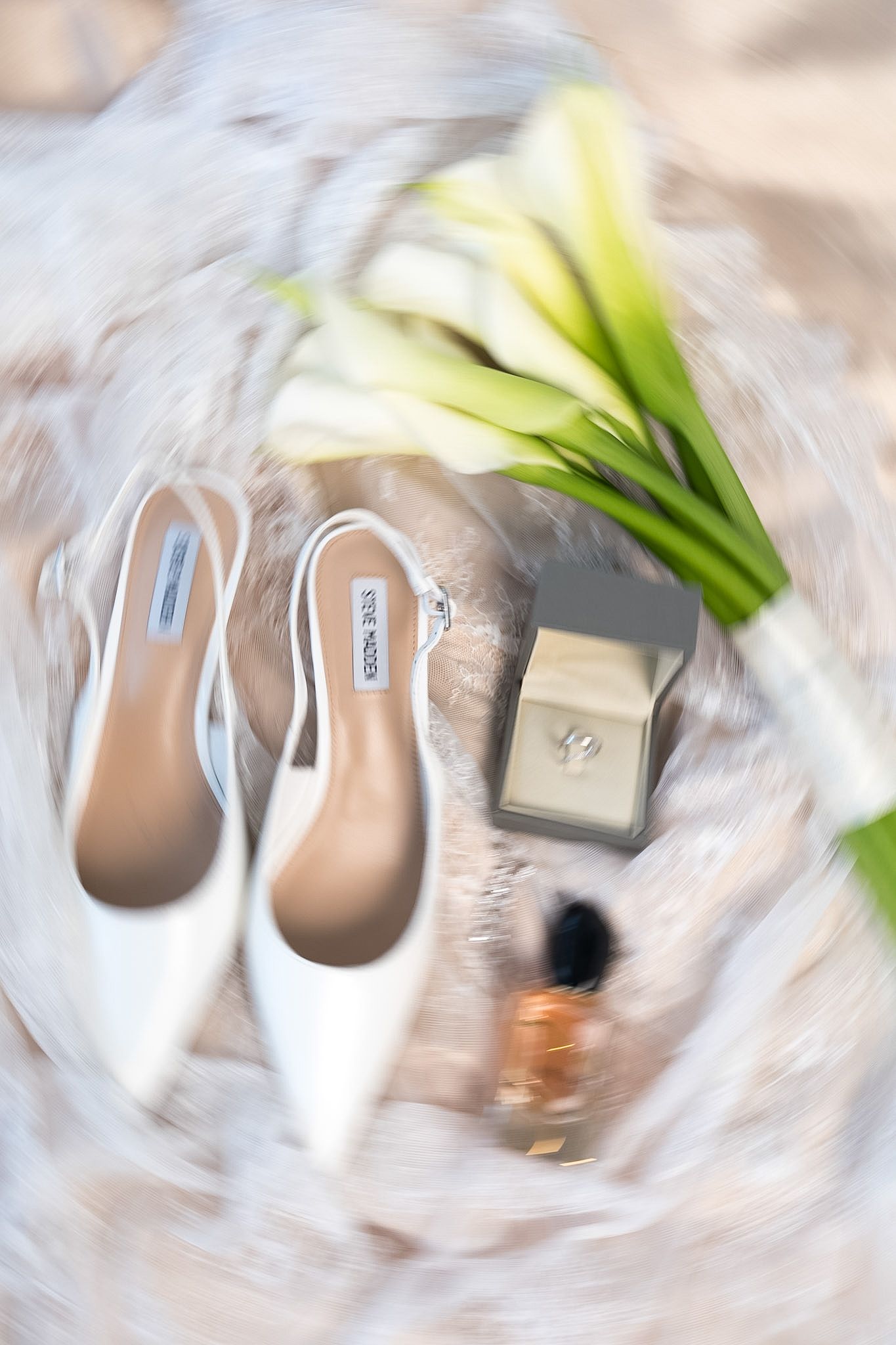 Flat lay of white pointed-toe heels, calla lily bouquet, ring box, and perfume on soft fabric.