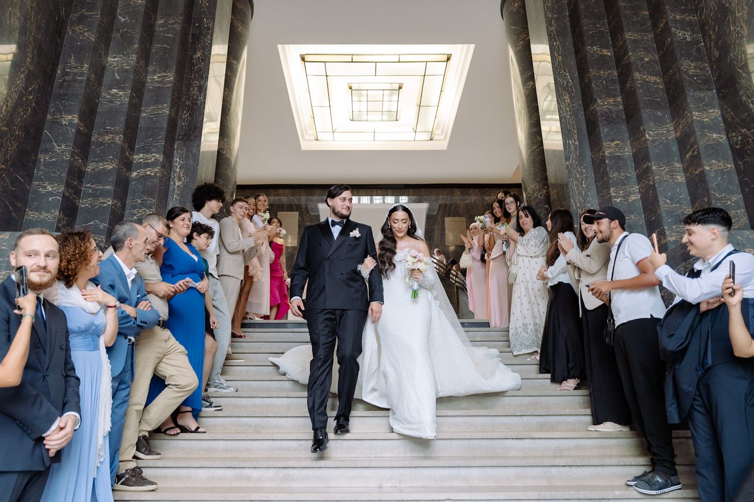 Bride and groom walk down grand marble staircase surrounded by cheering wedding guests in elegant venue.