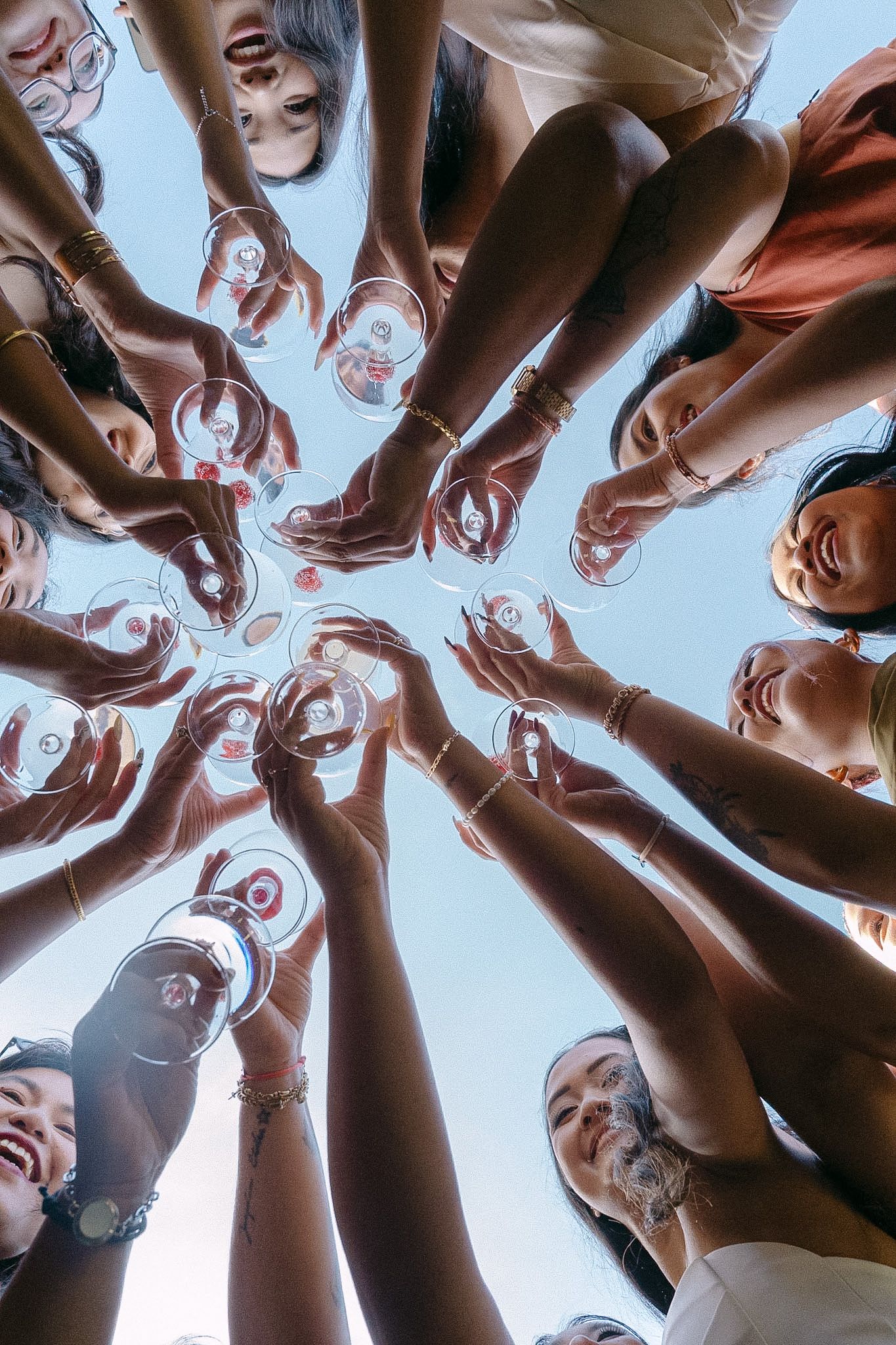 Group of women toasting with wine glasses in a circle, viewed from below against blue sky.