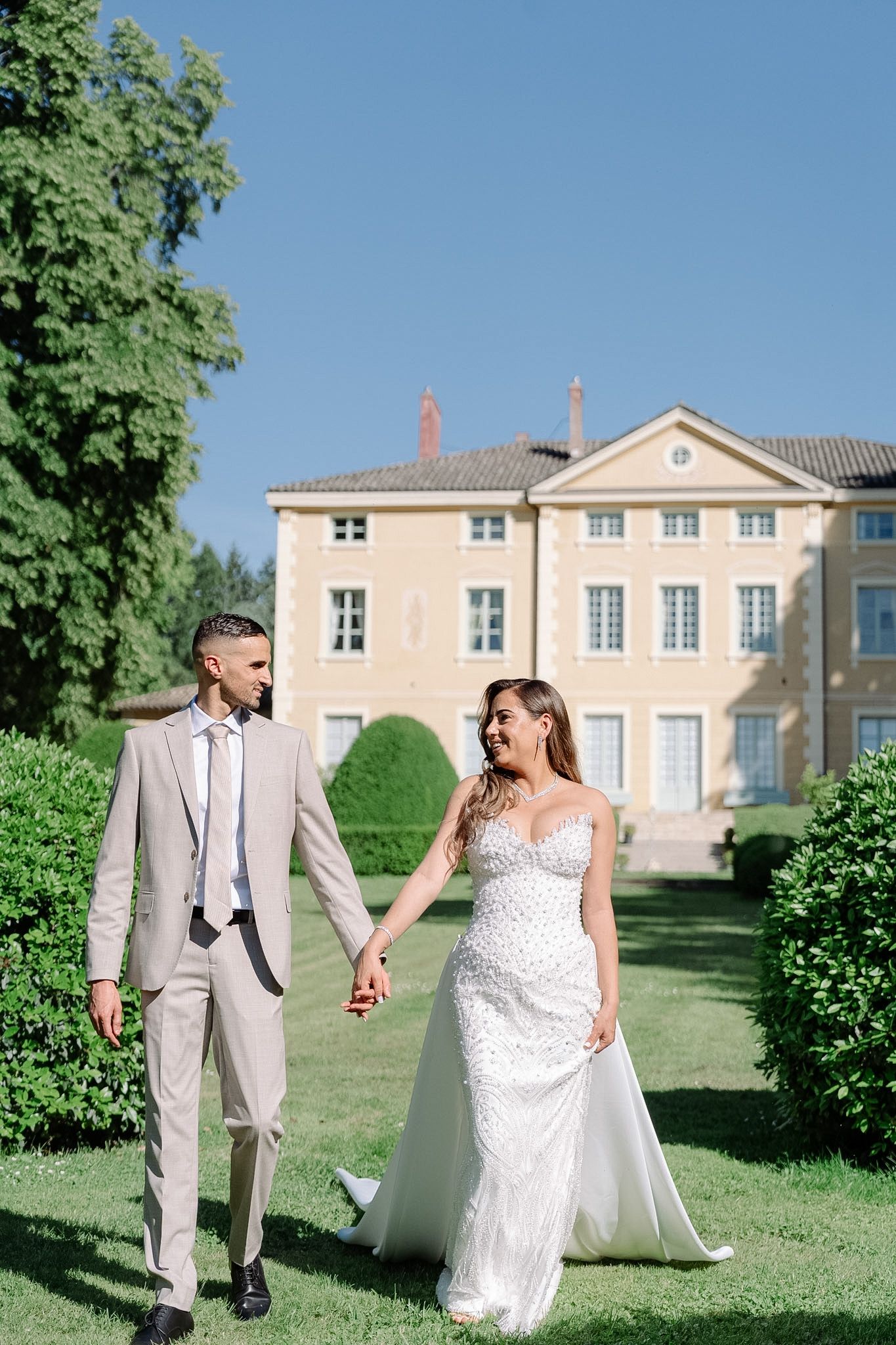 Bride and groom holding hands walking in lush garden with elegant French chateau in background on sunny day.