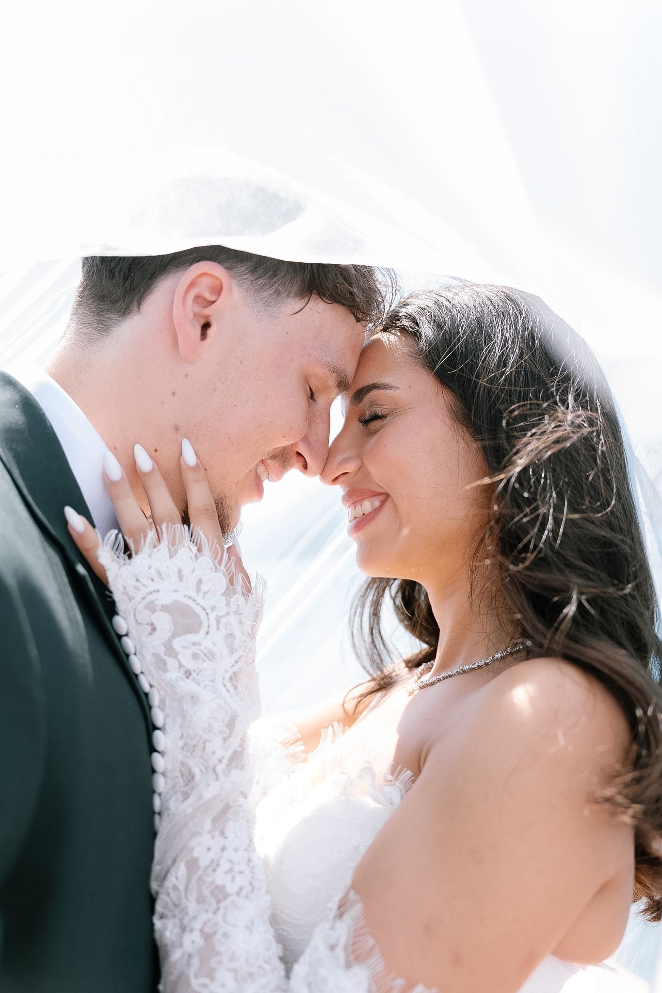 A bride and groom share an intimate forehead touch under a flowing white veil on their wedding day.
