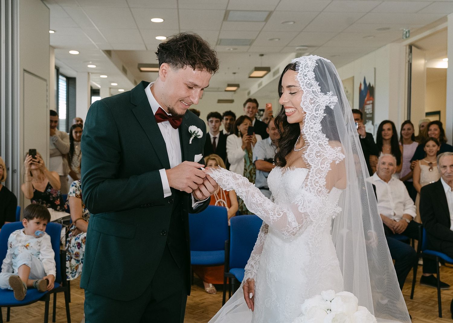 Groom in green suit places ring on bride in lace wedding dress and veil during indoor ceremony.