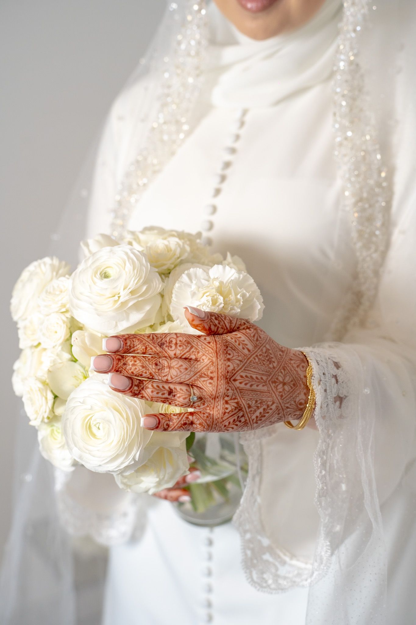 A bride in a white dress holds a white rose bouquet while displaying intricate henna designs on her hands.