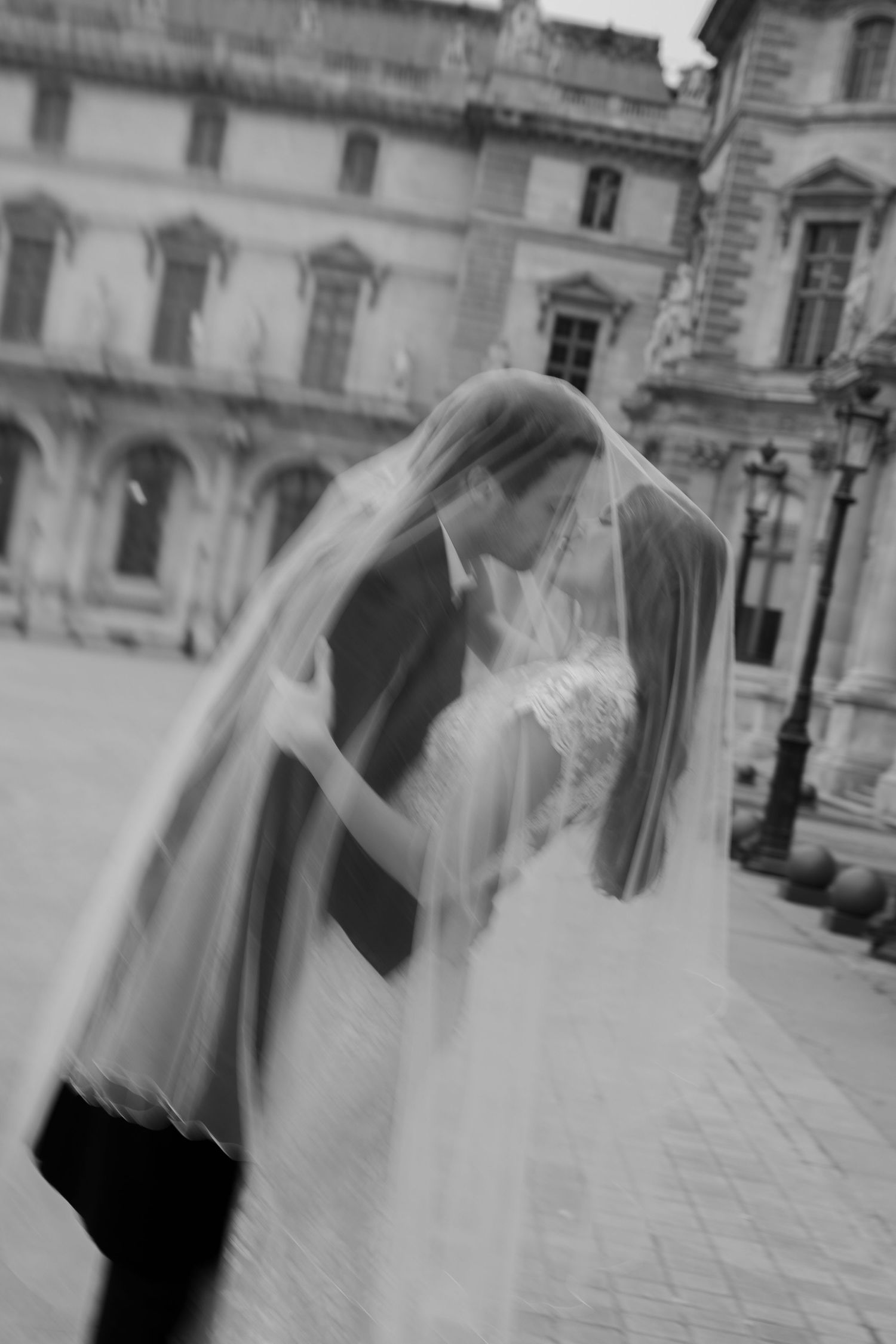 Black and white photo of a bride and groom kissing under a veil outside a grand building.