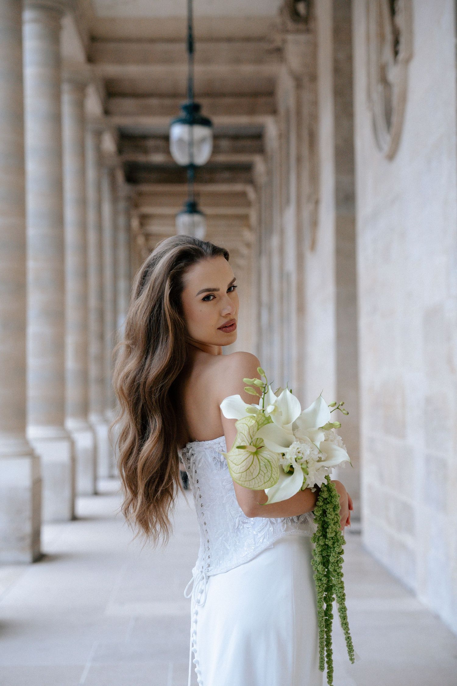 Bride in white off-shoulder corset gown holding white lily bouquet in elegant stone colonnade.