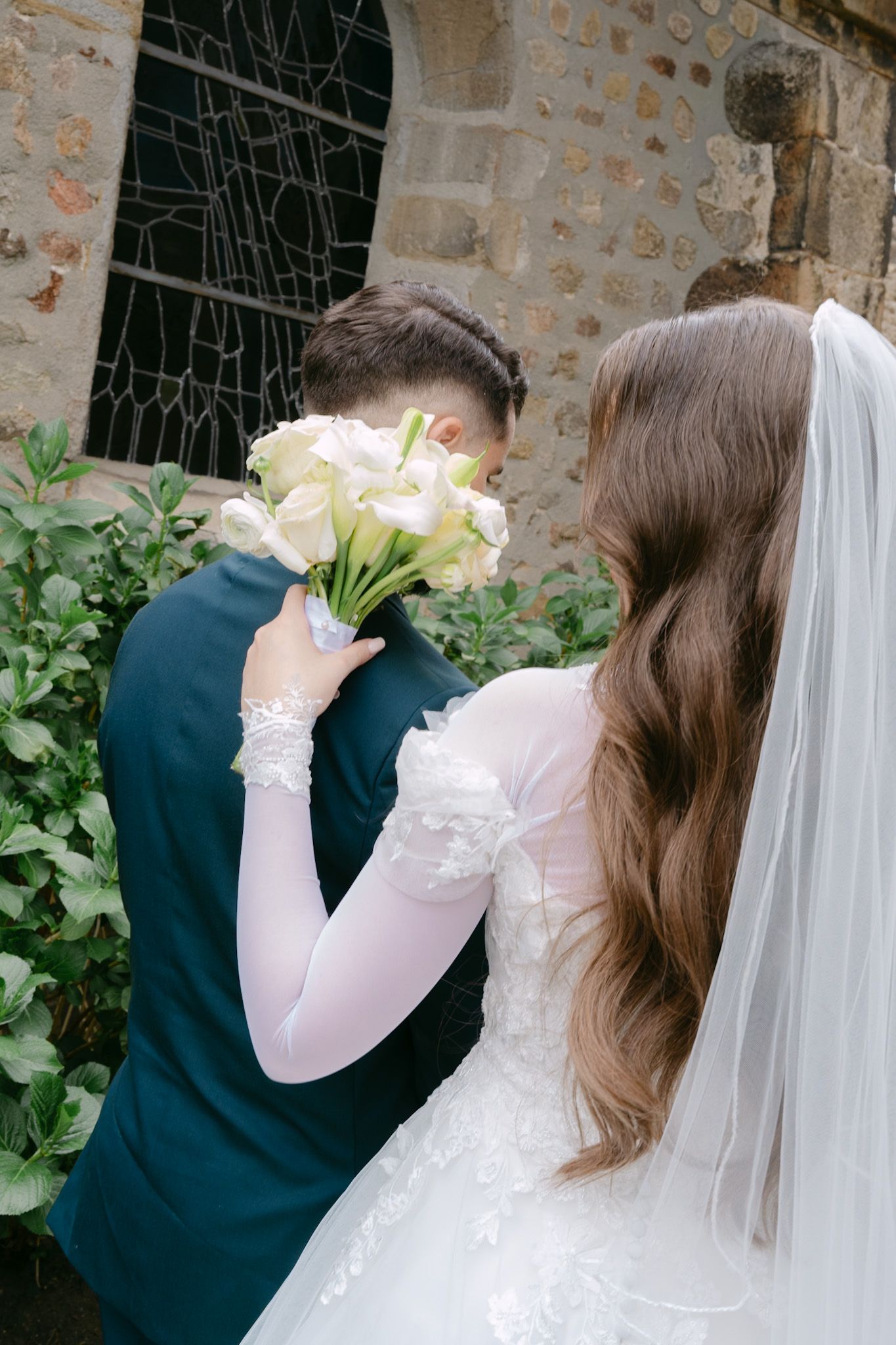 Bride and groom kissing behind white calla lily bouquet outdoors near stone wall on wedding day.