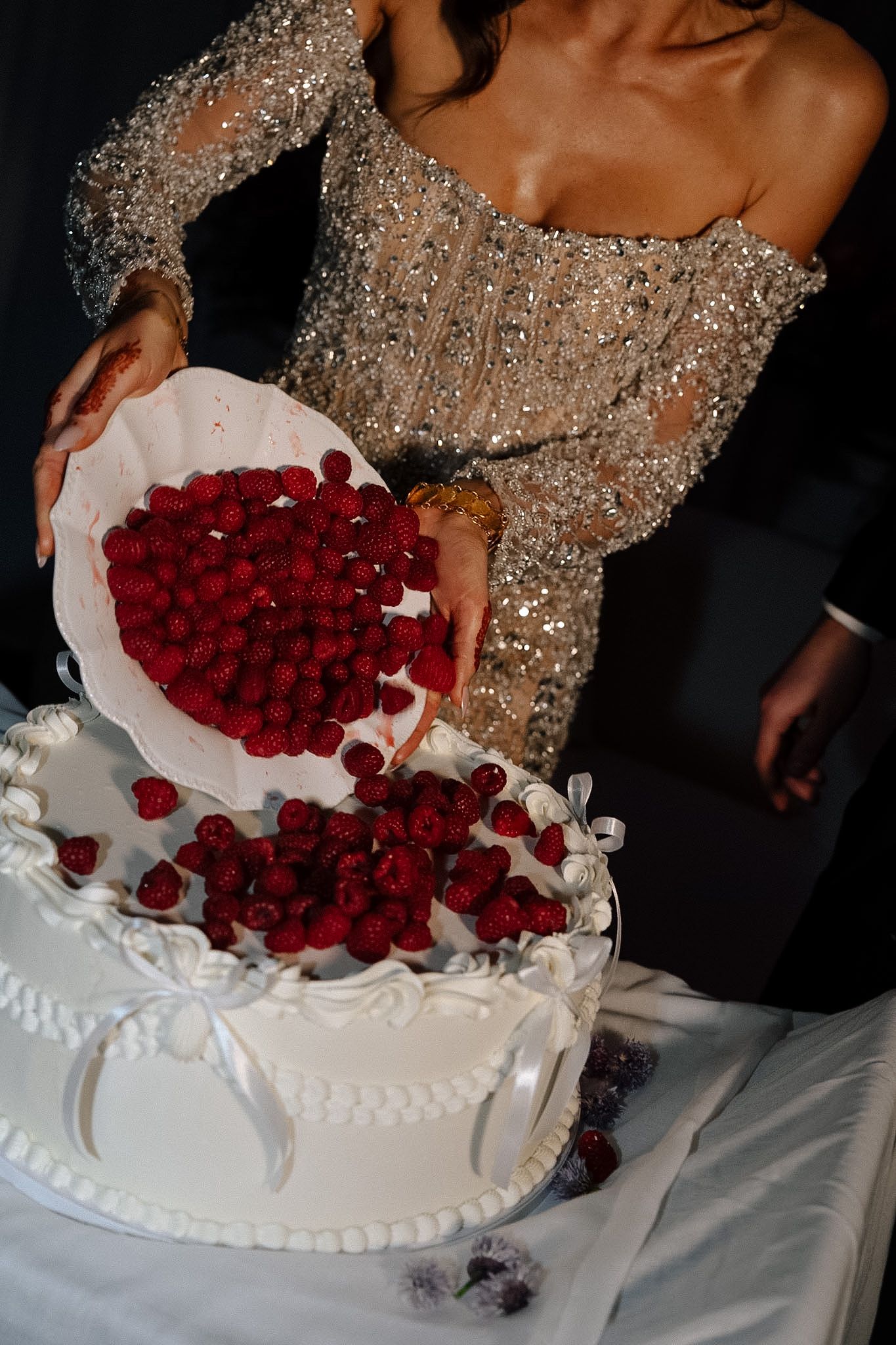Woman in silver sequin gown pouring fresh raspberries onto elegant white wedding cake.