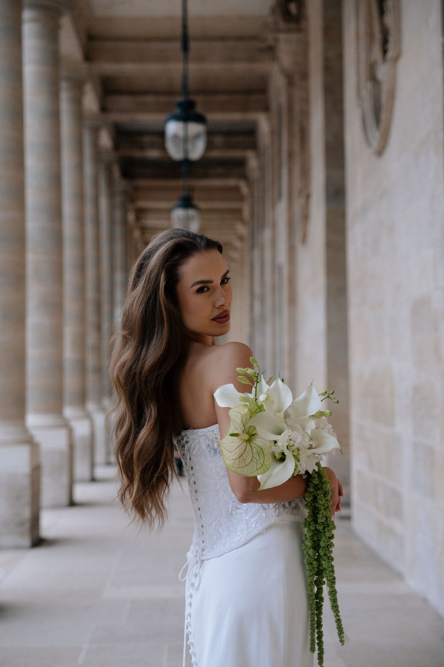 Bride in white off-shoulder gown holding white lily bouquet in elegant stone colonnade.