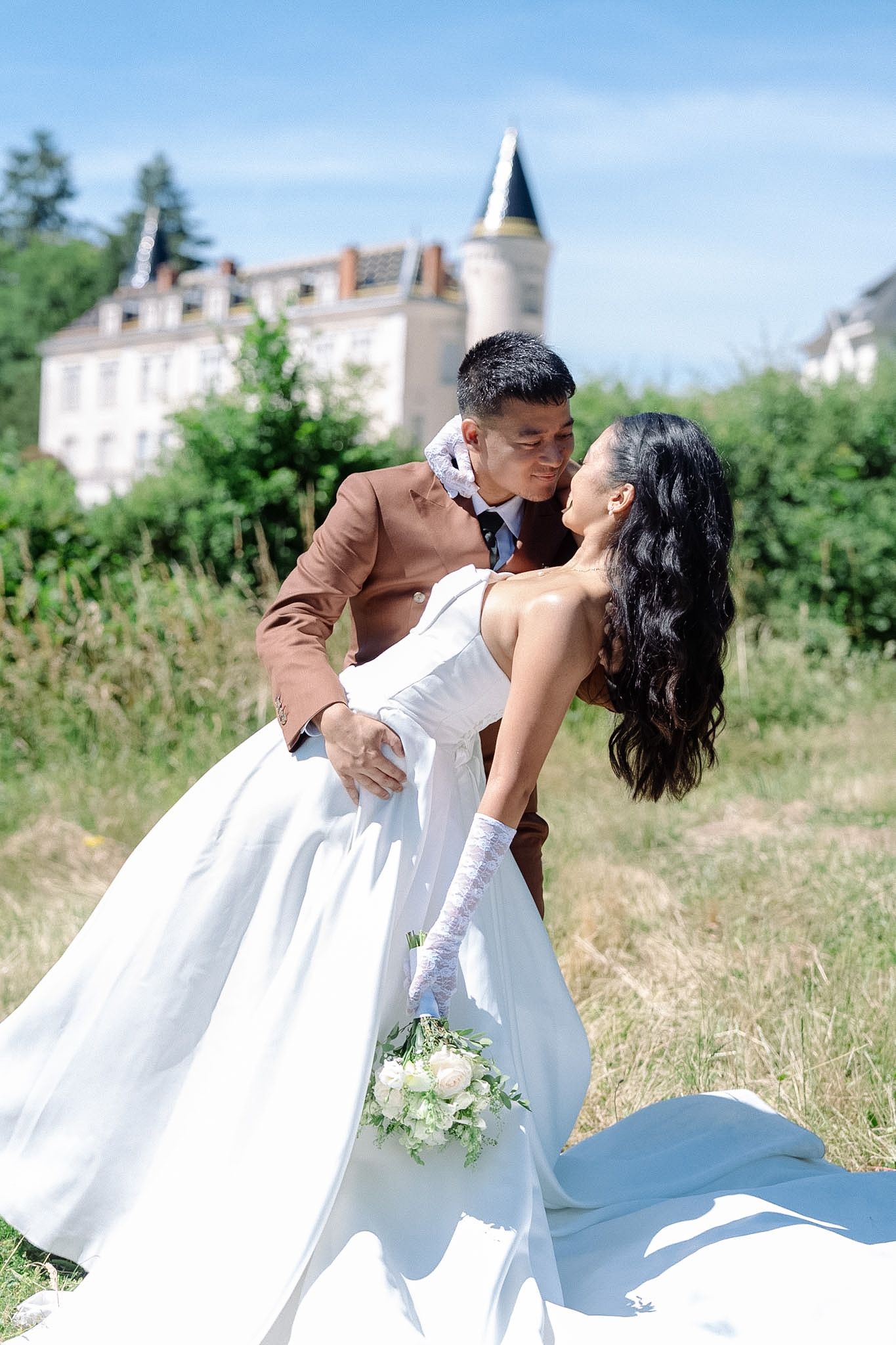 Bride and groom share a romantic dip kiss outdoors with a French château and blue sky in the background.