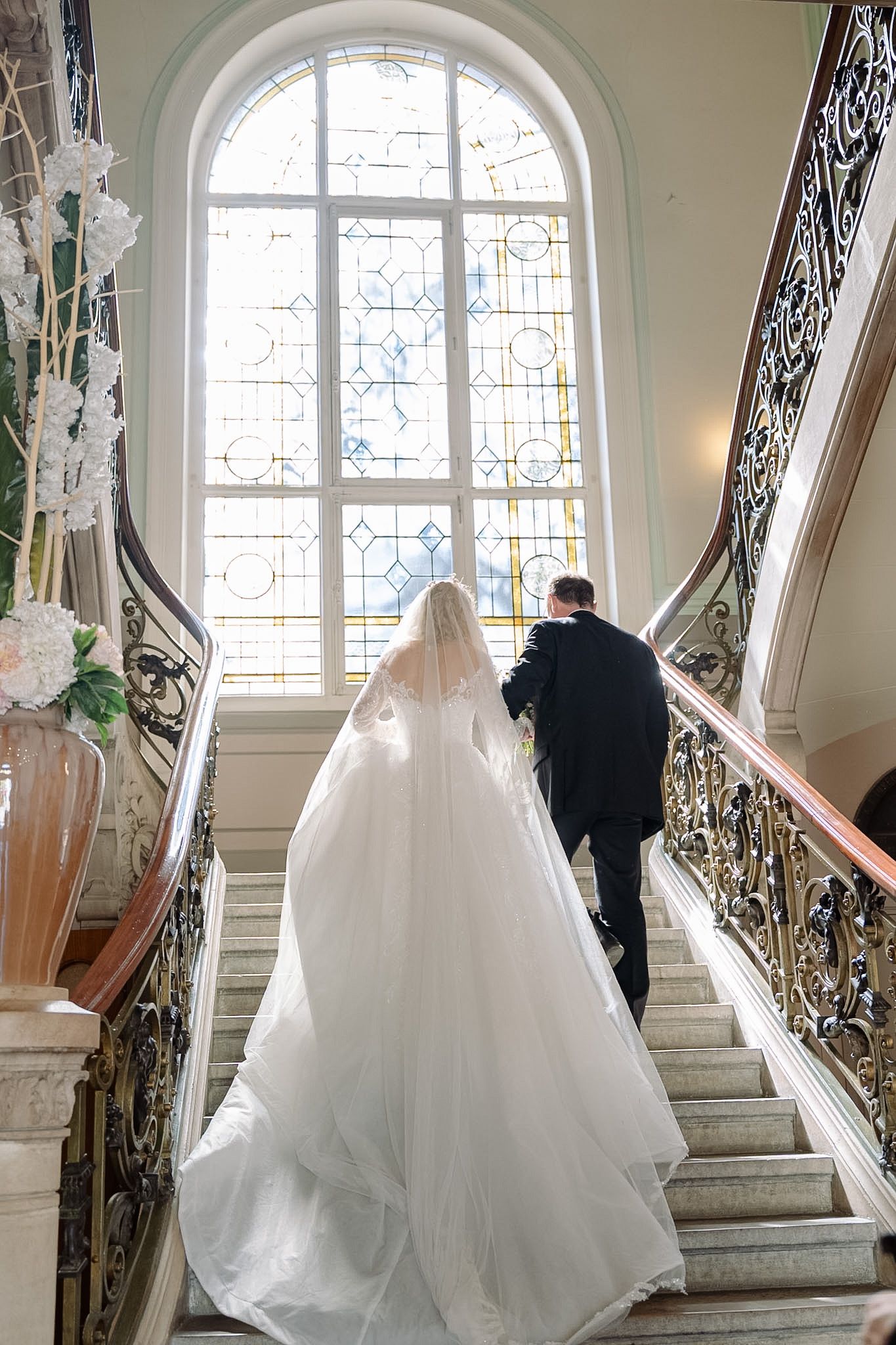 Bride and groom ascending elegant staircase with ornate railings and arched stained glass window.
