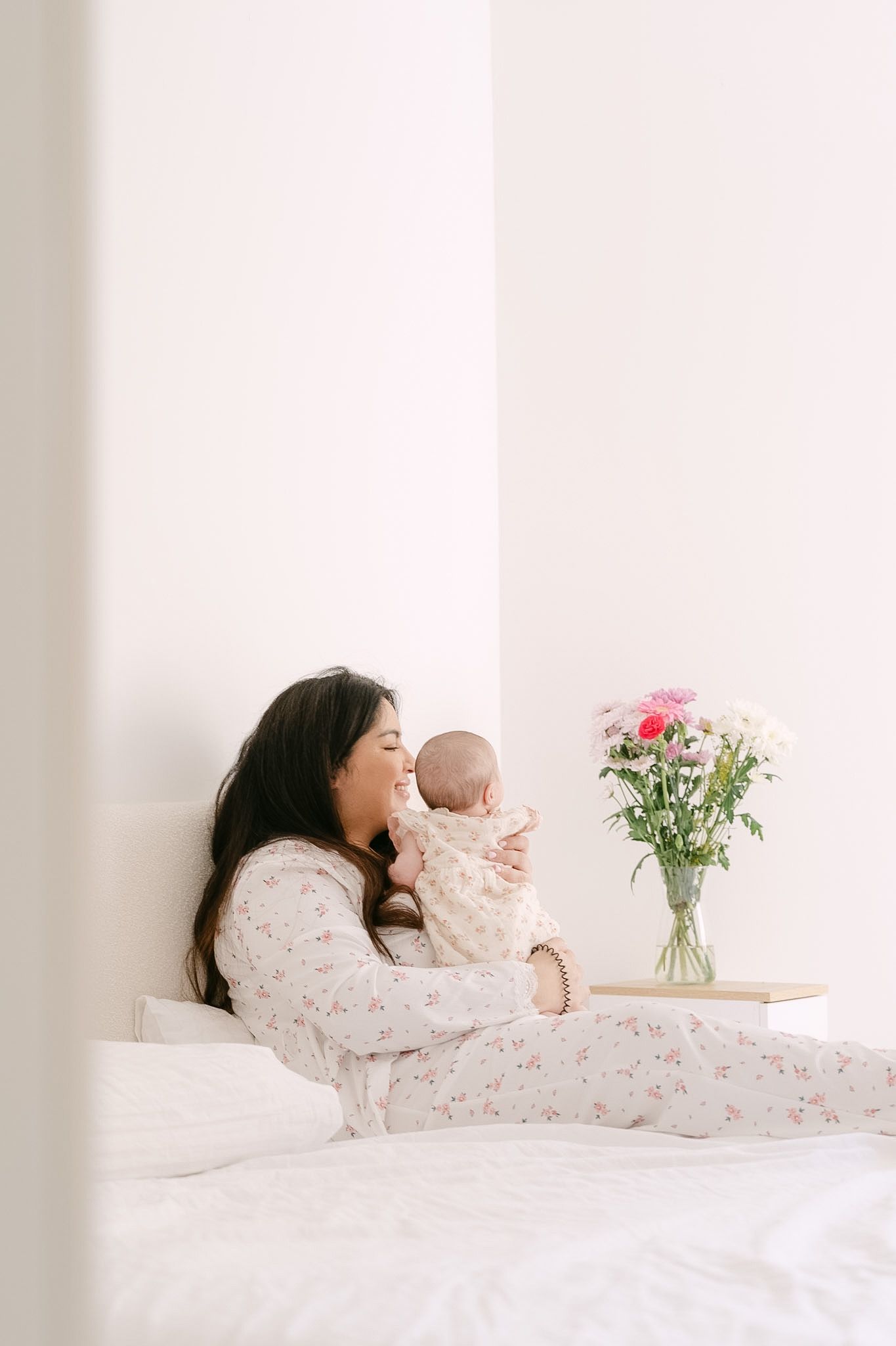 A mother in floral pajamas sits on a white bed, lovingly holding her newborn baby near a vase of pink flowers.