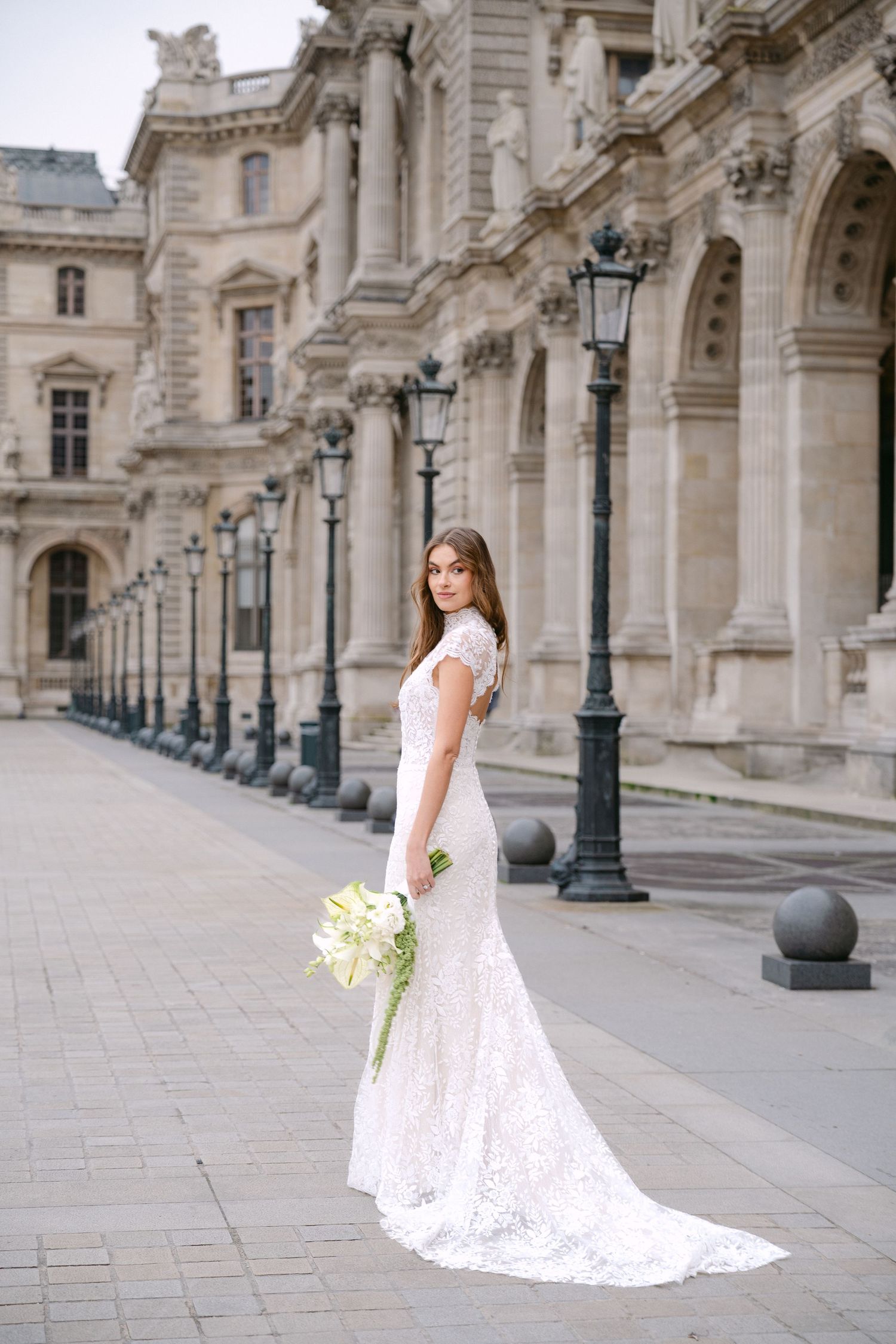 Bride in white lace gown holding bouquet outside the Louvre in Paris, France.