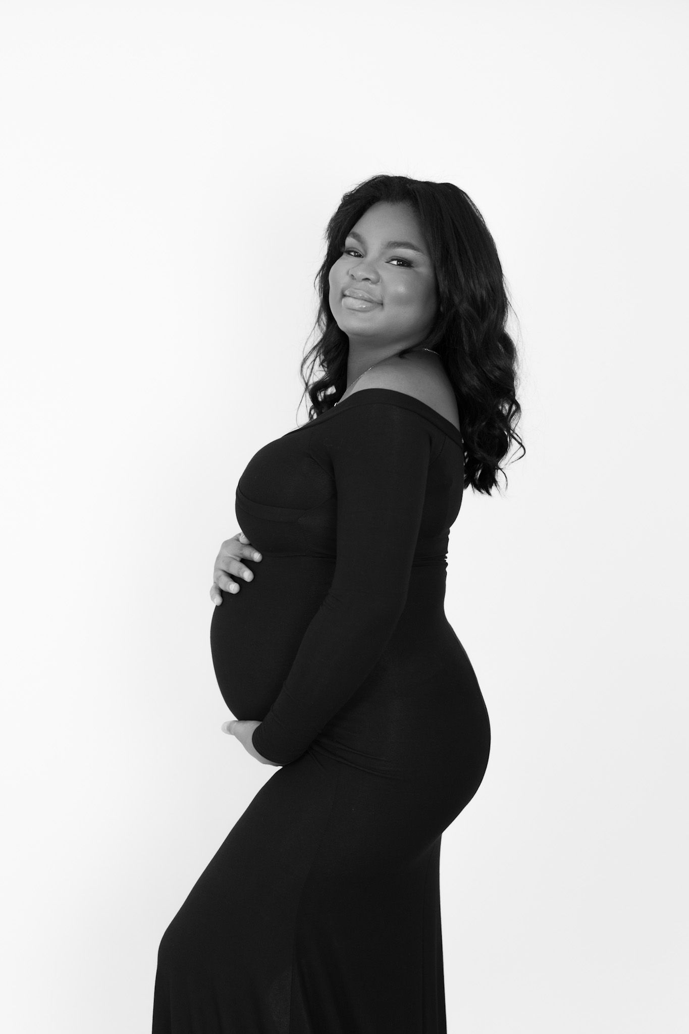 Black and white maternity photo of a smiling pregnant woman in a black off-shoulder gown cradling her baby bump.