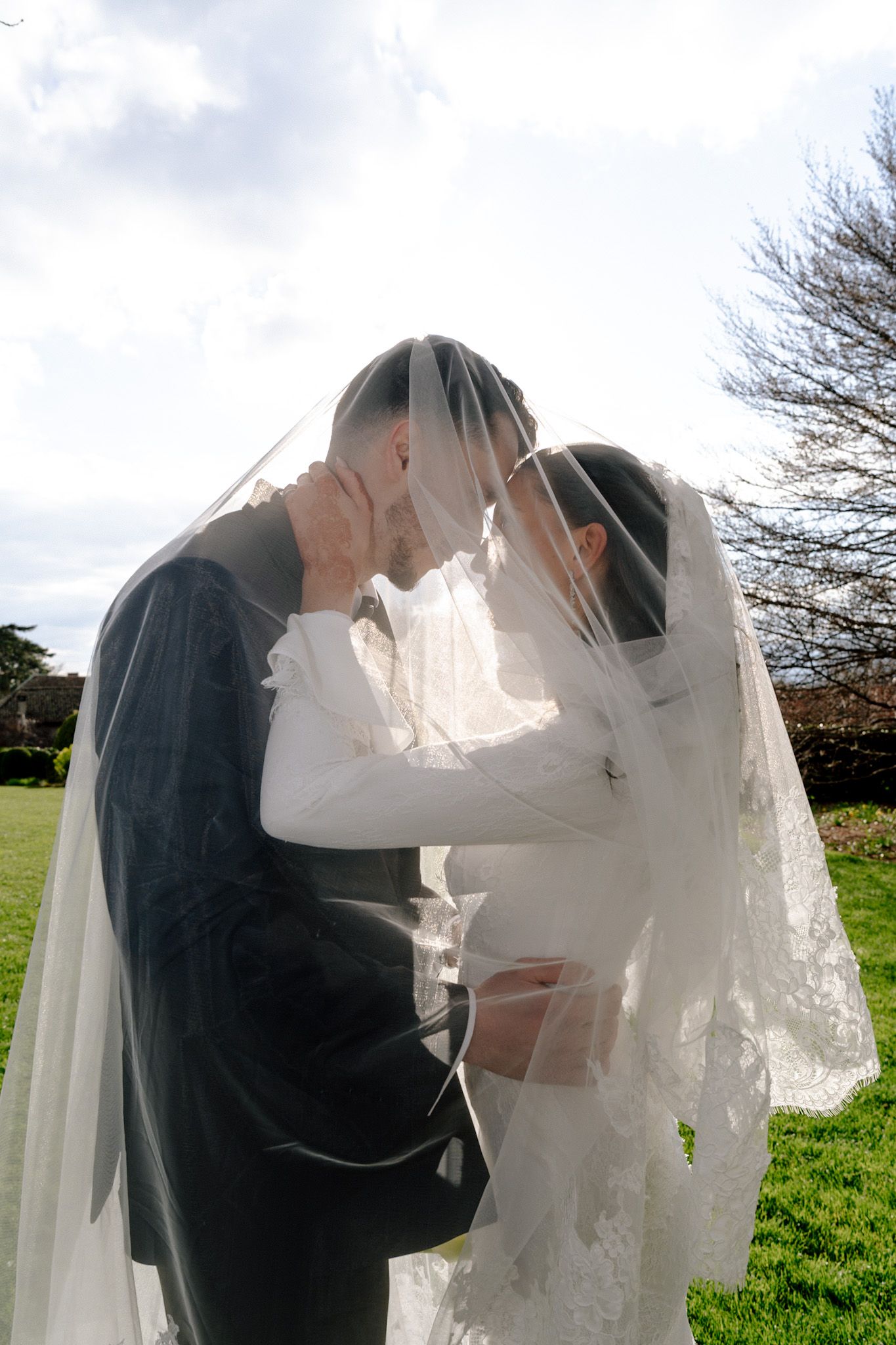 Bride and groom sharing an intimate moment under a flowing white wedding veil outdoors on a green lawn.
