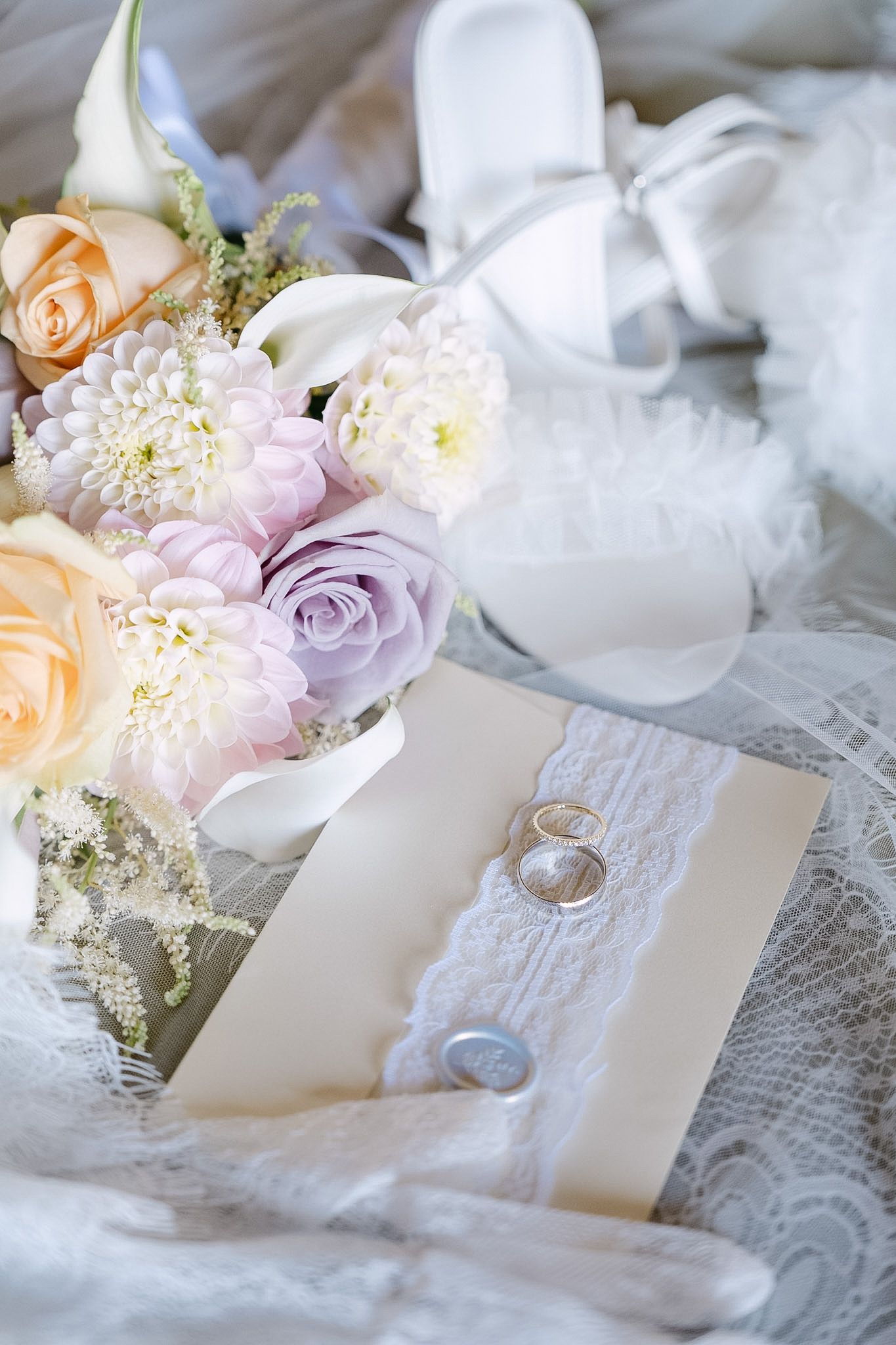Elegant wedding flat lay with pastel bouquet, white heels, lace veil, rings, and ivory invitation card.