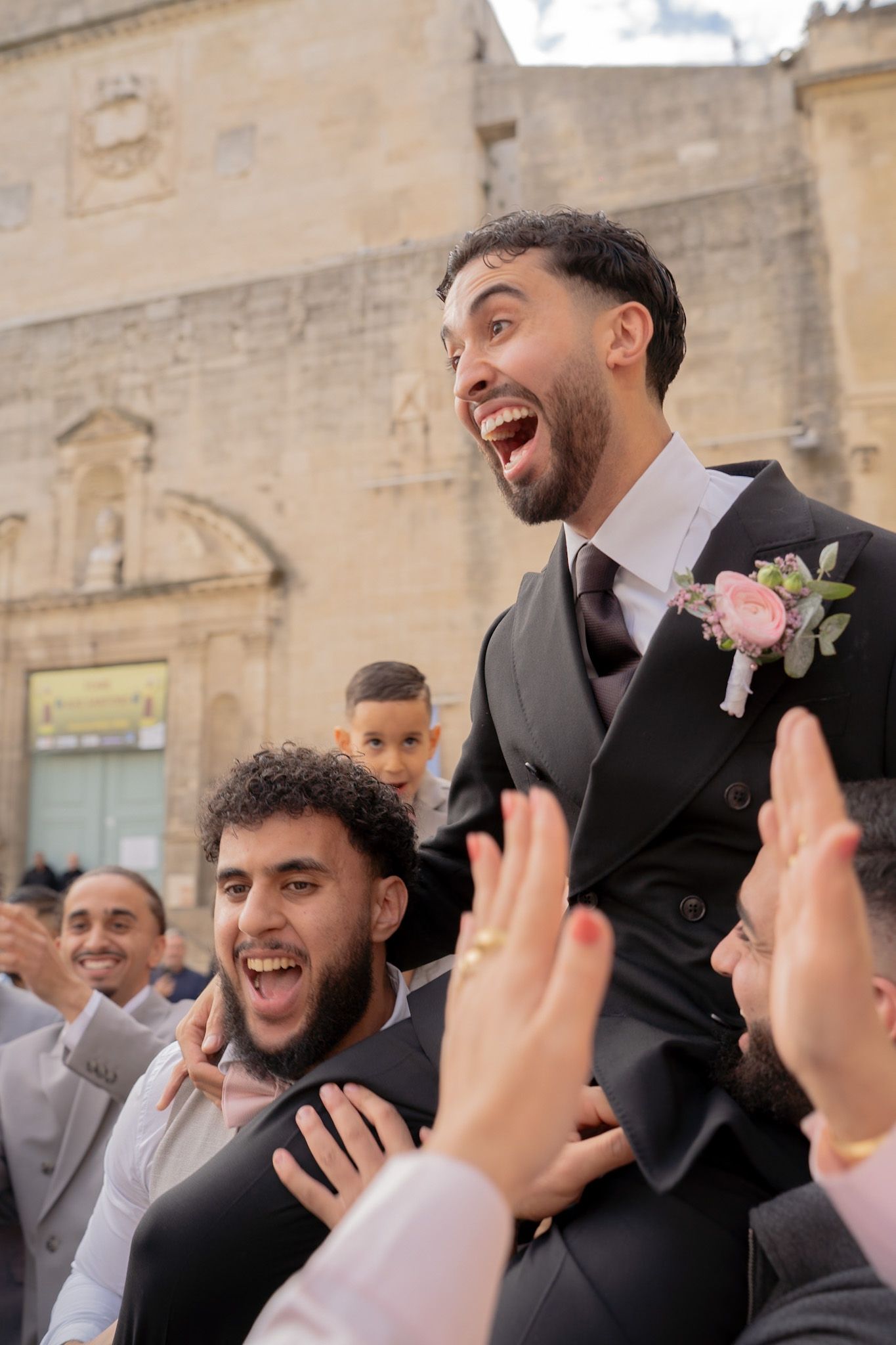 Joyful groom in black suit with pink boutonniere lifted by friends during outdoor wedding celebration.