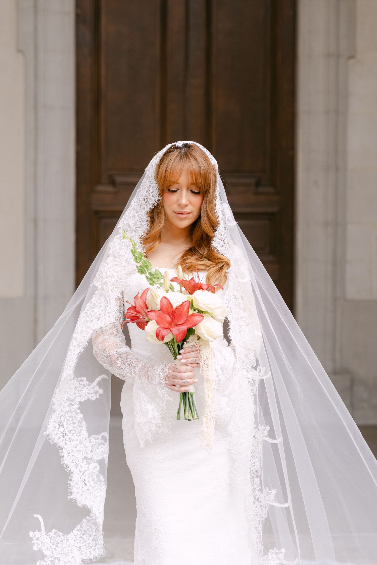 Elegant bride in lace wedding dress and veil holding red and white flower bouquet outside church doors.