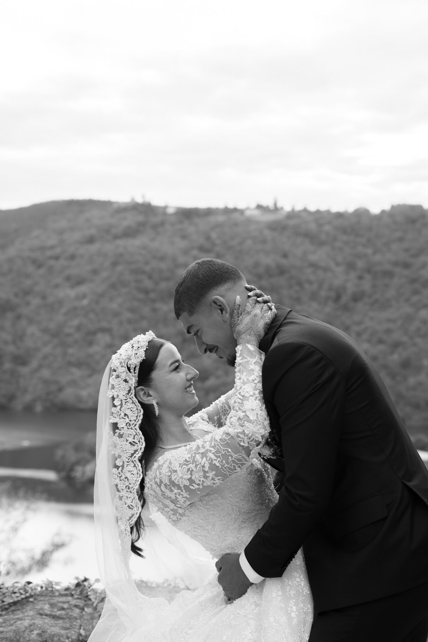 A romantic black and white wedding photo of a couple embracing outdoors with mountains and water in the background.