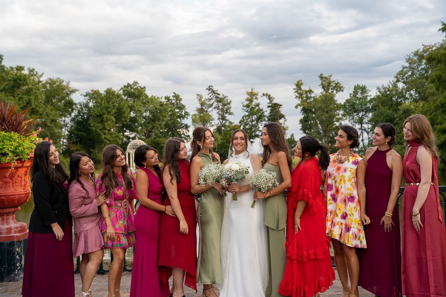 Bride in white gown surrounded by bridesmaids in colorful red and pink dresses, laughing outdoors at a wedding celebration.