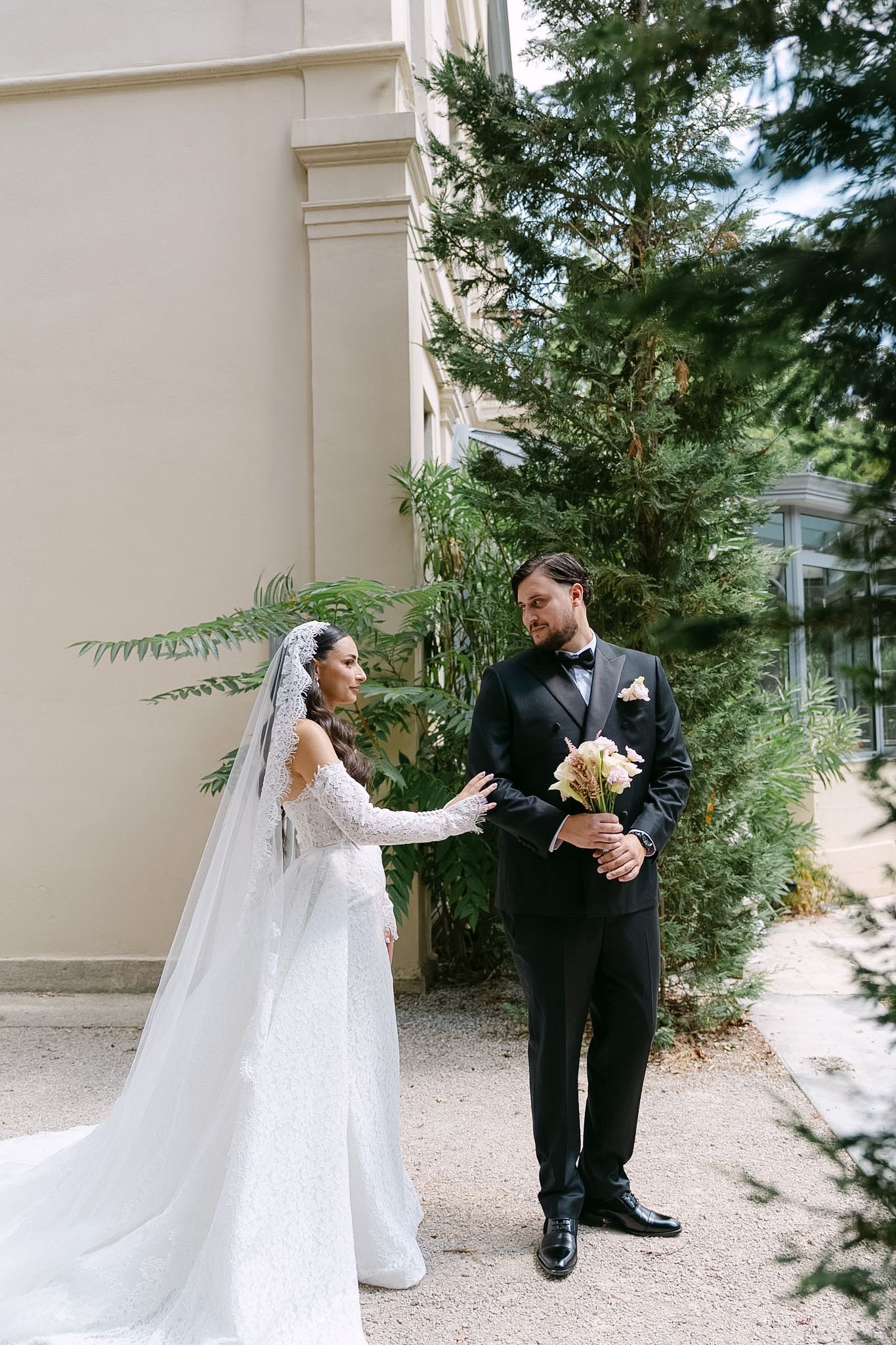 Bride in white lace gown and veil hands bouquet to groom in black tuxedo outdoors near evergreen trees.