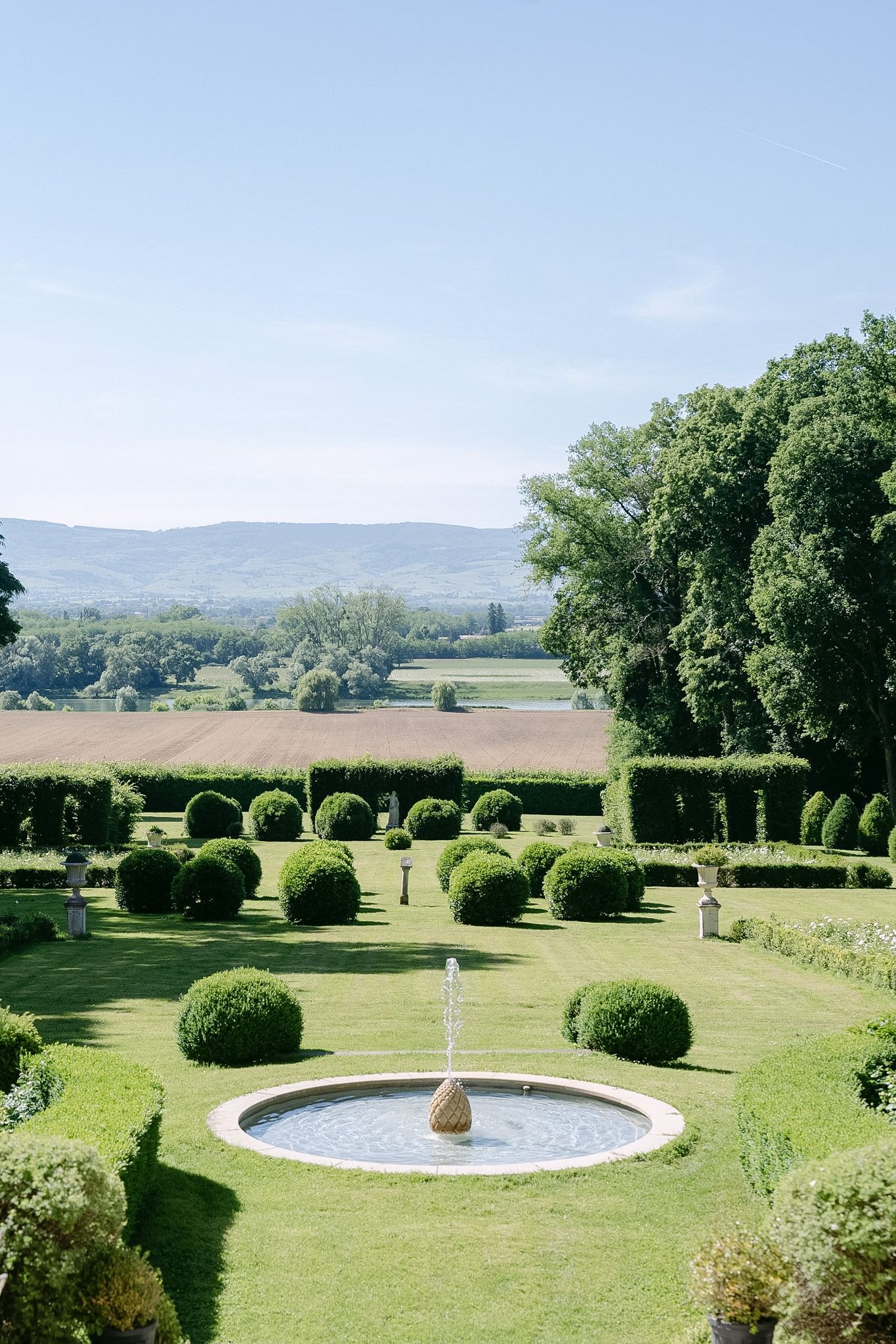 Elegant formal garden with circular fountain, manicured round topiary bushes, green lawn, and scenic mountain views under clear blue sky.