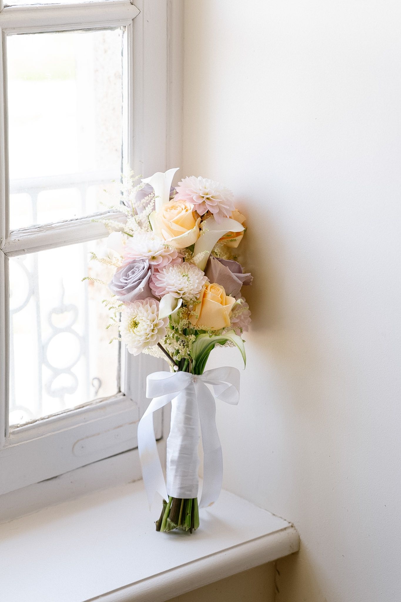 Elegant bridal bouquet of pastel roses and white flowers tied with ribbon, resting on windowsill in soft natural light.