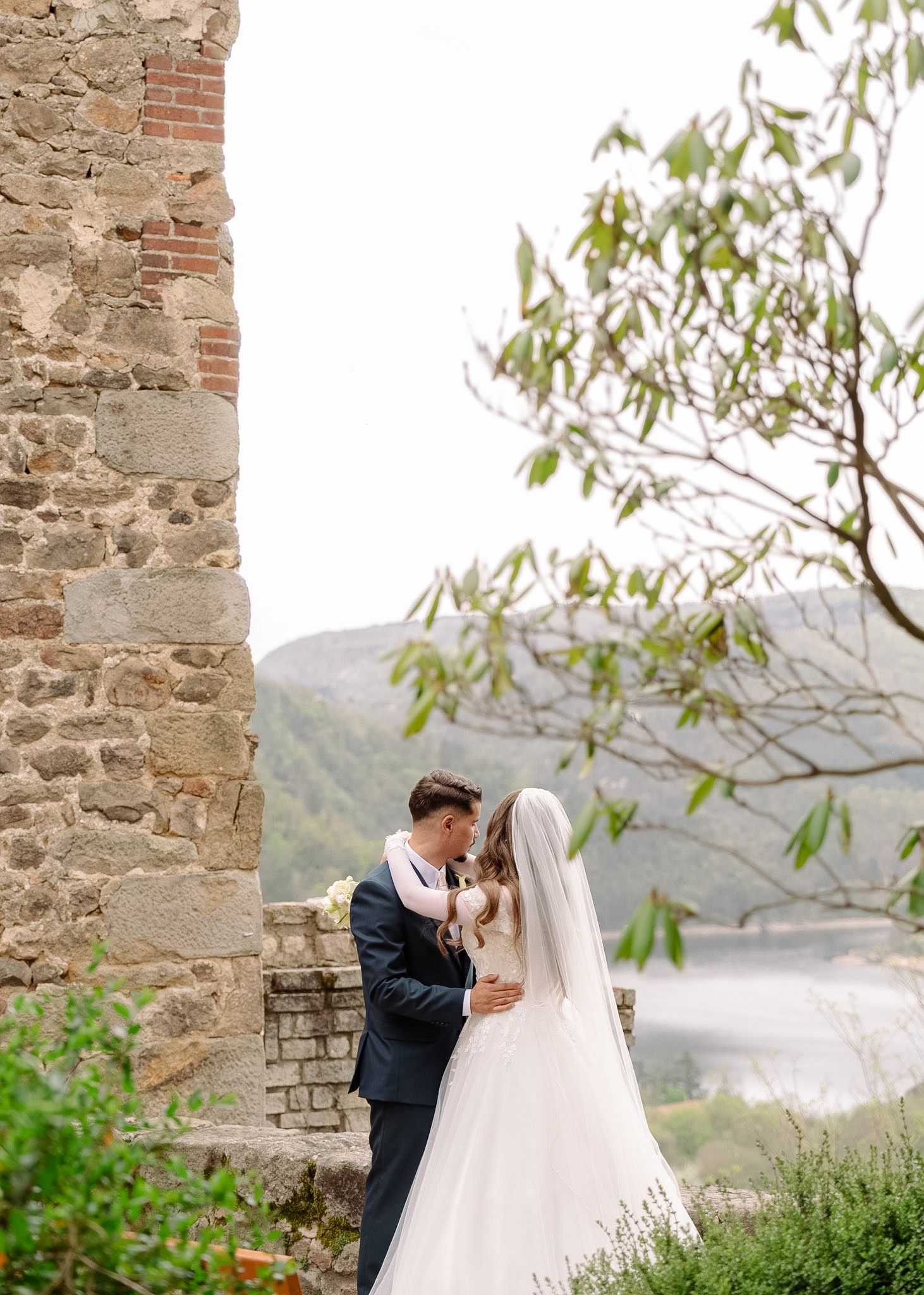 Bride and groom share a romantic kiss beside a stone castle wall with scenic mountain lake views.