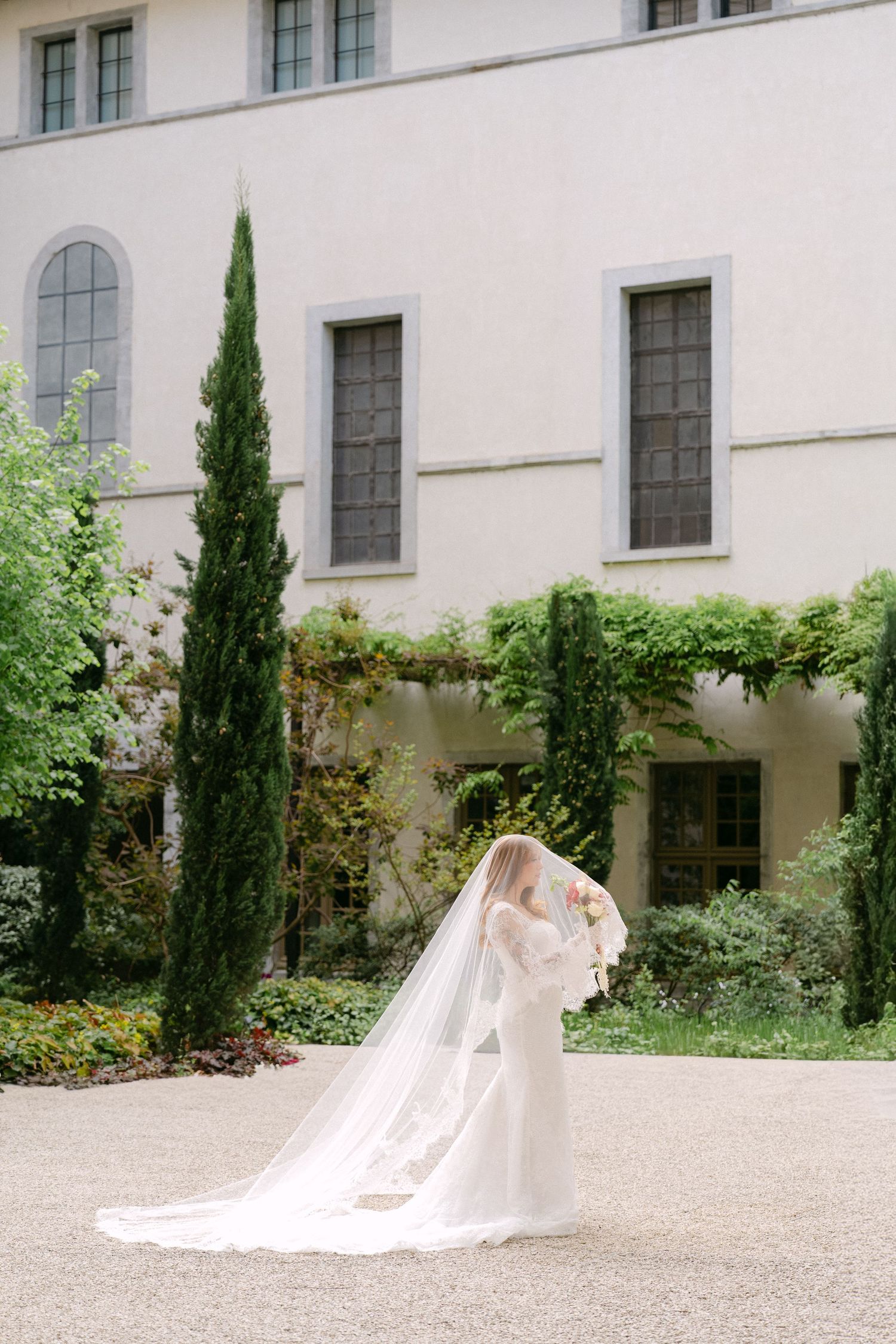 Bride in a flowing white gown and long veil stands in an elegant garden courtyard with tall cypress trees.