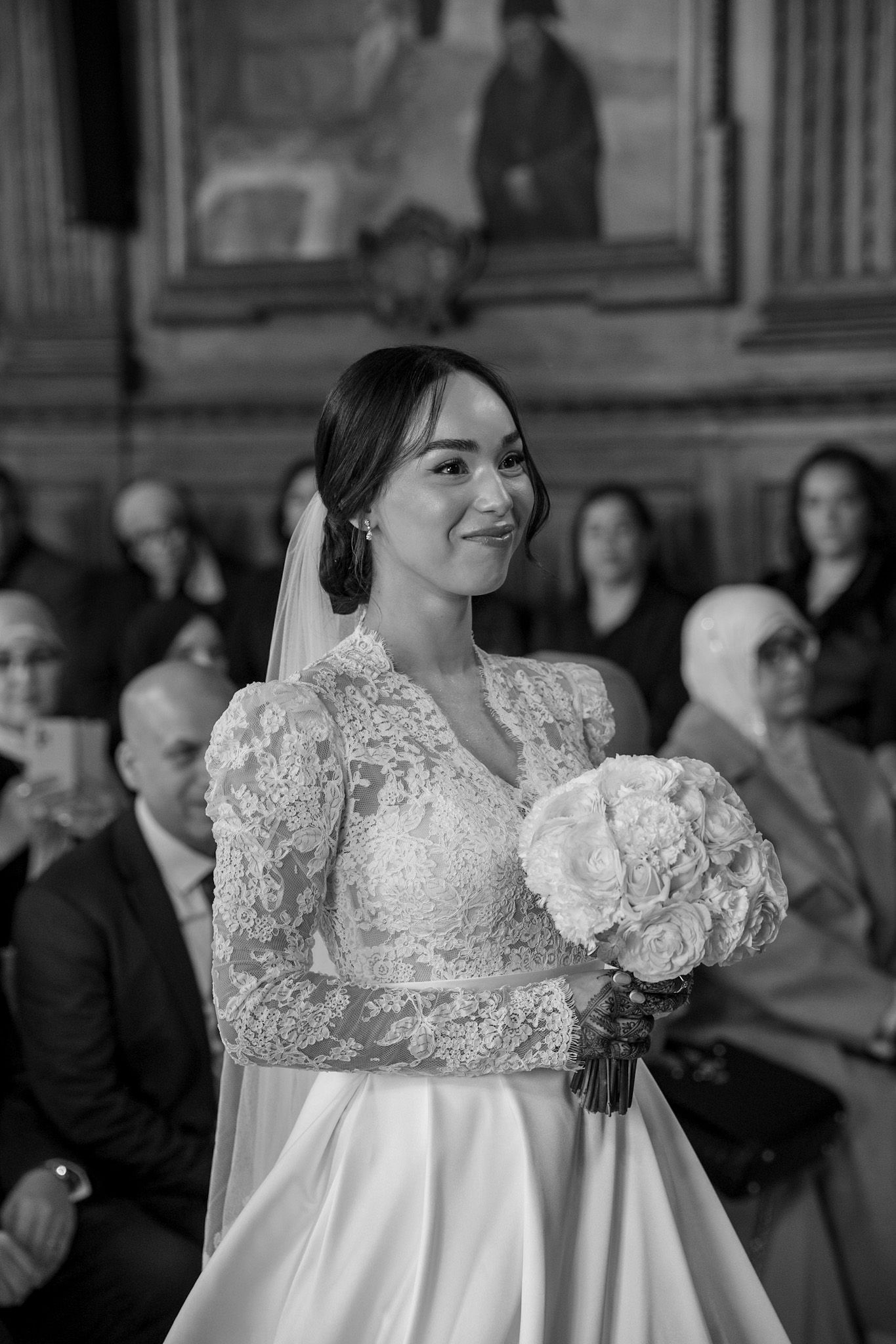 Black and white photo of smiling bride in lace wedding gown holding bouquet during ceremony.