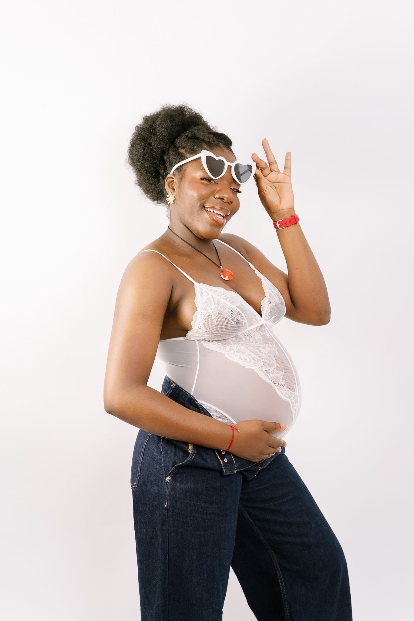 Smiling pregnant woman in white lace top and jeans, wearing heart-shaped sunglasses, posing against white background.