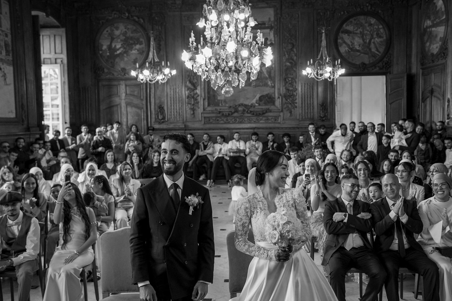 Black and white wedding ceremony photo in an ornate hall with chandeliers, guests seated as couple stands at altar.