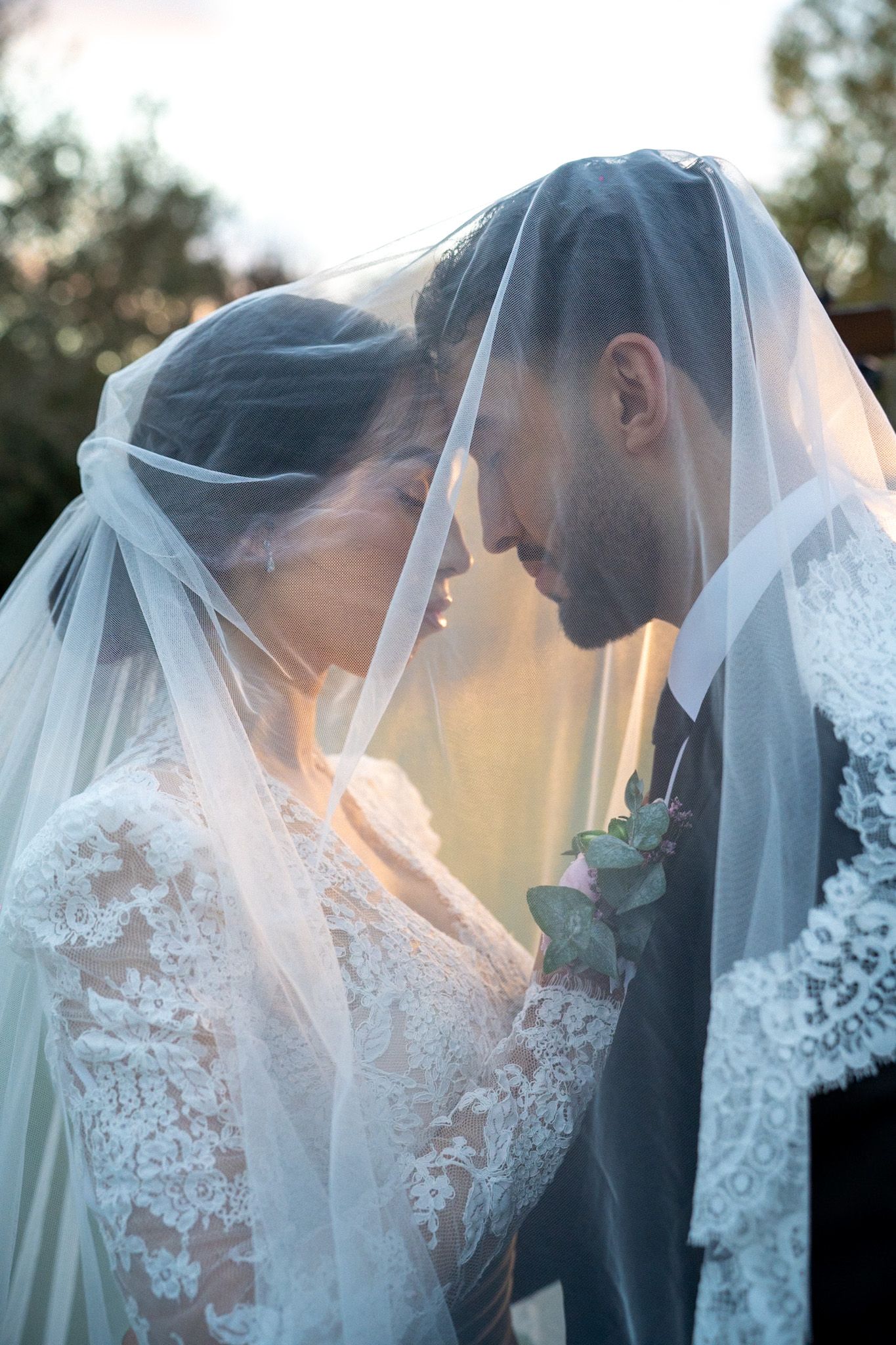 Bride and groom share an intimate moment under a wedding veil, foreheads touching in a romantic outdoor setting.