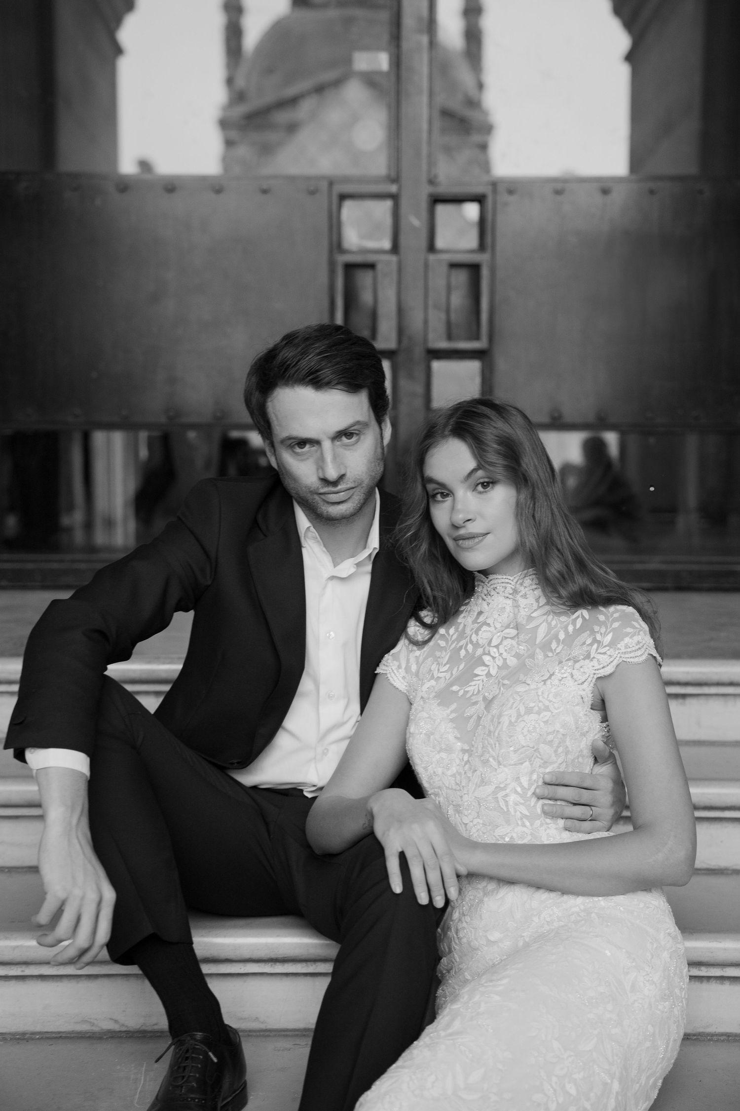 Black and white photo of an elegant couple in wedding attire sitting together on stone steps.