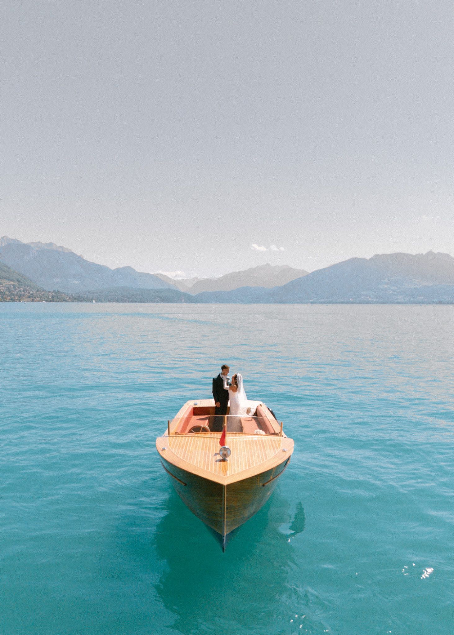 Couple embracing on a wooden boat on turquoise alpine lake with mountain backdrop on their wedding day.