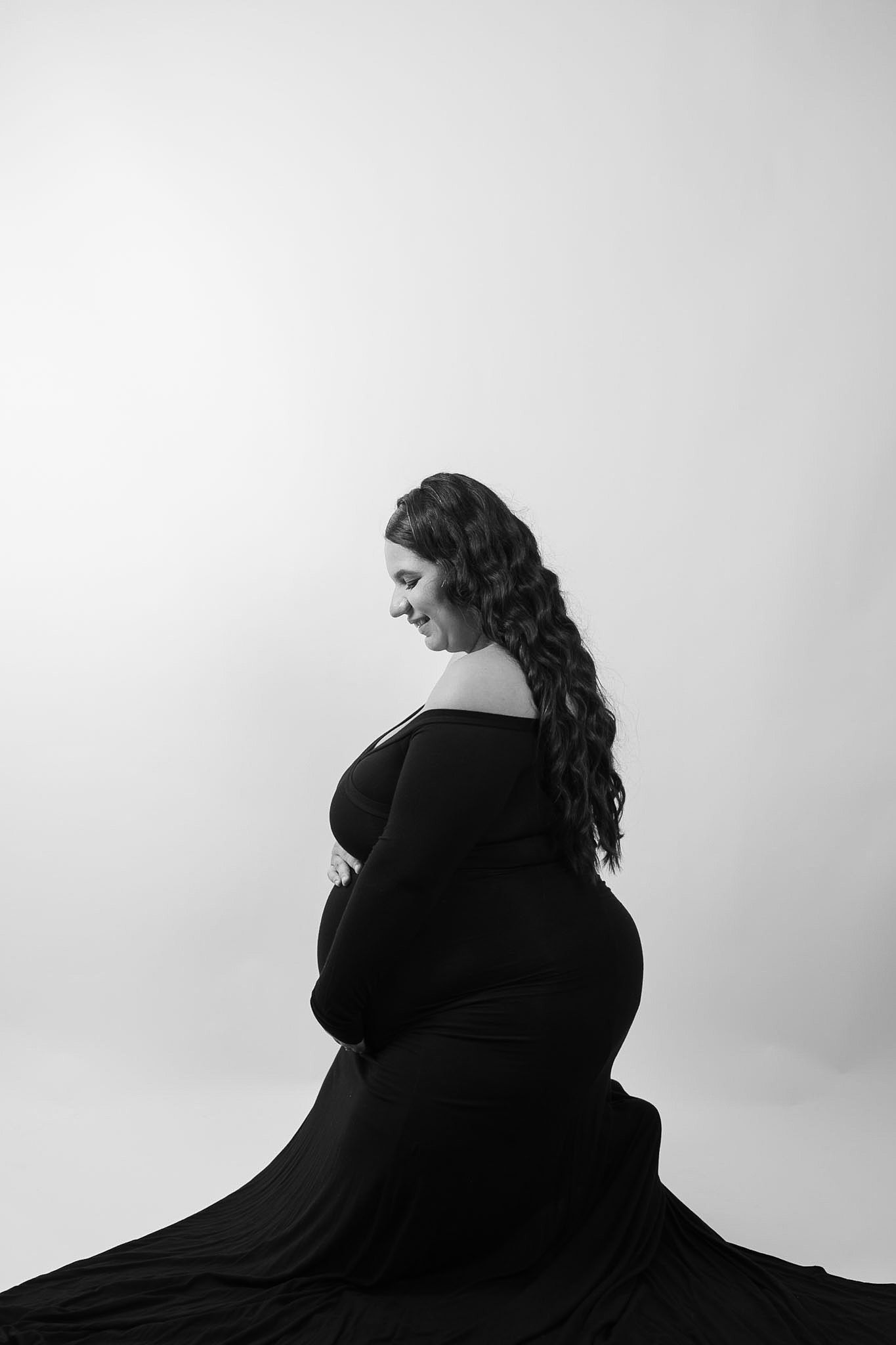 Black and white maternity photo of a pregnant woman in a flowing black gown, sitting gracefully in a studio.